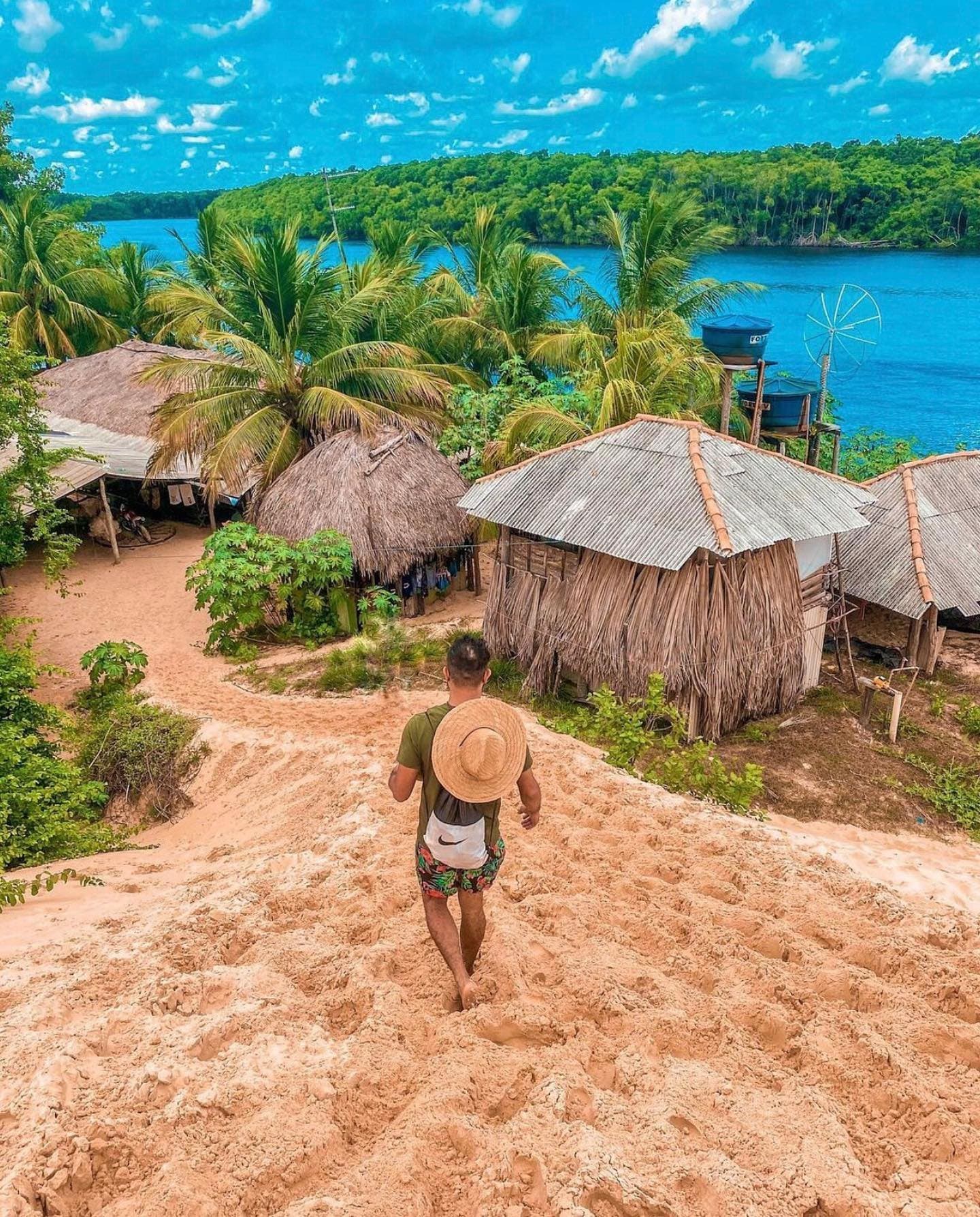 Some villages you walk into, and some you slide into. This one qualifies. A traveller with a straw hat and backpack walks barefoot down a sandy slope toward thatched and tin-roofed huts nestled among palm trees beside a wide blue river on the Lençóis Maranhenses full itinerary