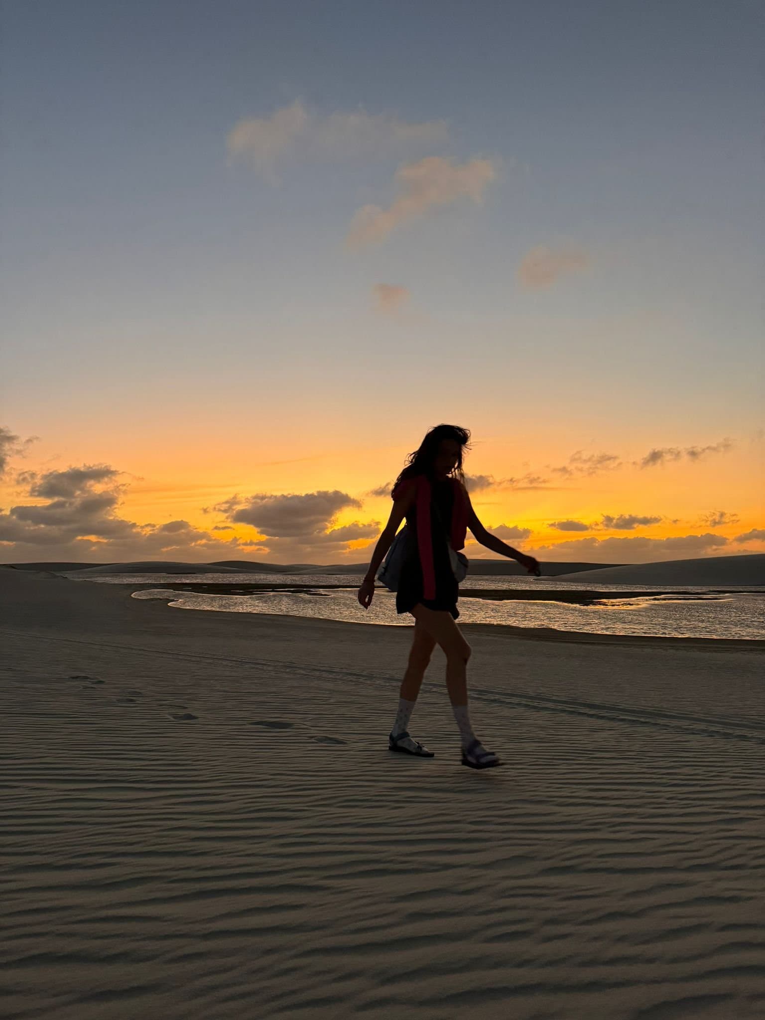Your own footprints in the sand, the light doing the rest. A woman's silhouette walks across rippled sand dunes at golden-hour sunset on the Lençóis Maranhenses full itinerary, with a glimmering lagoon and rolling dunes stretching to the horizon behind her