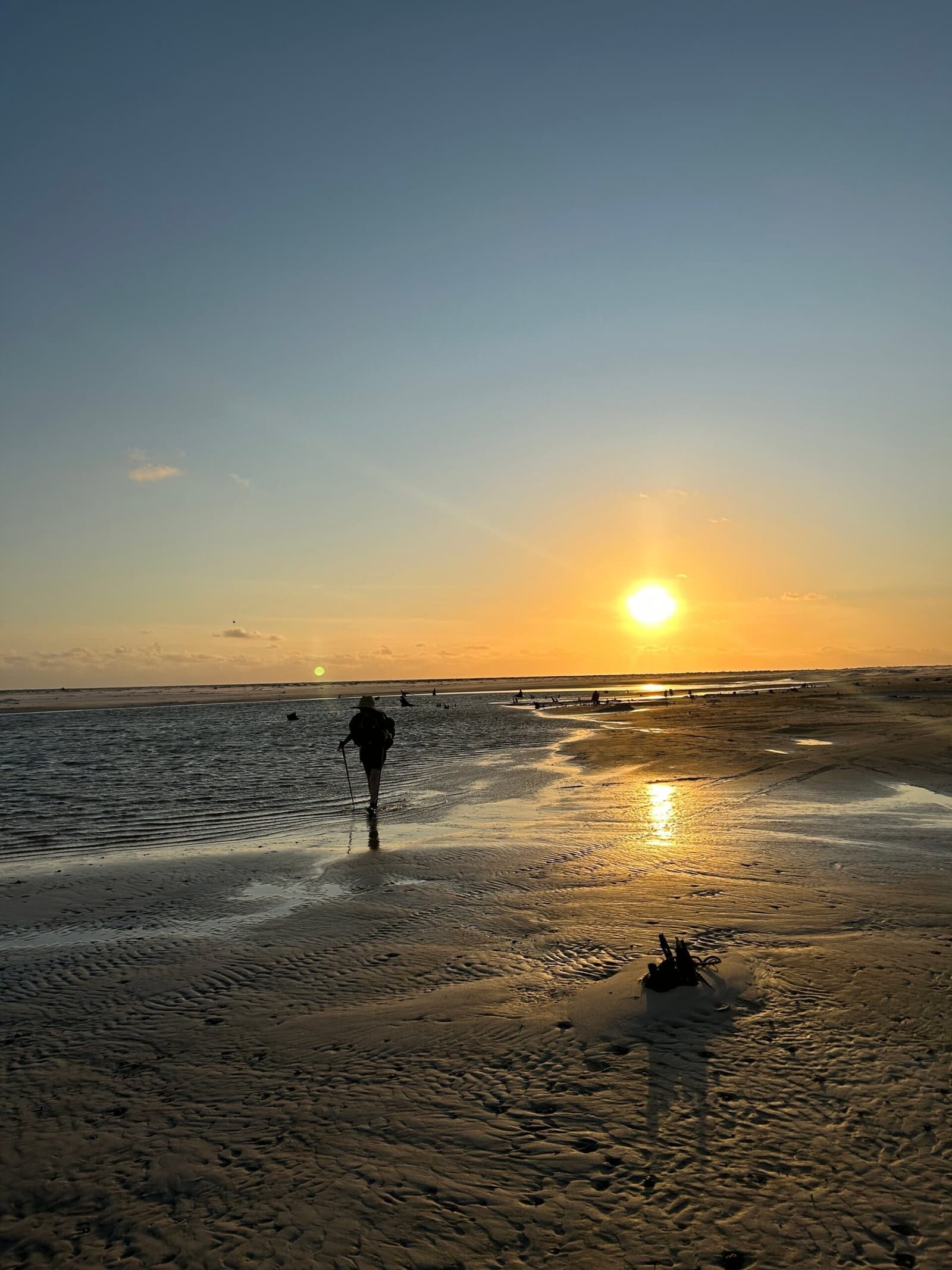 One more step toward the light, the flats have a way of making you feel small and free. A lone hiker with trekking poles wades through a shallow tidal channel on the Lençóis Maranhenses hike, silhouetted against a golden sunset over the flat, rippled sandflats