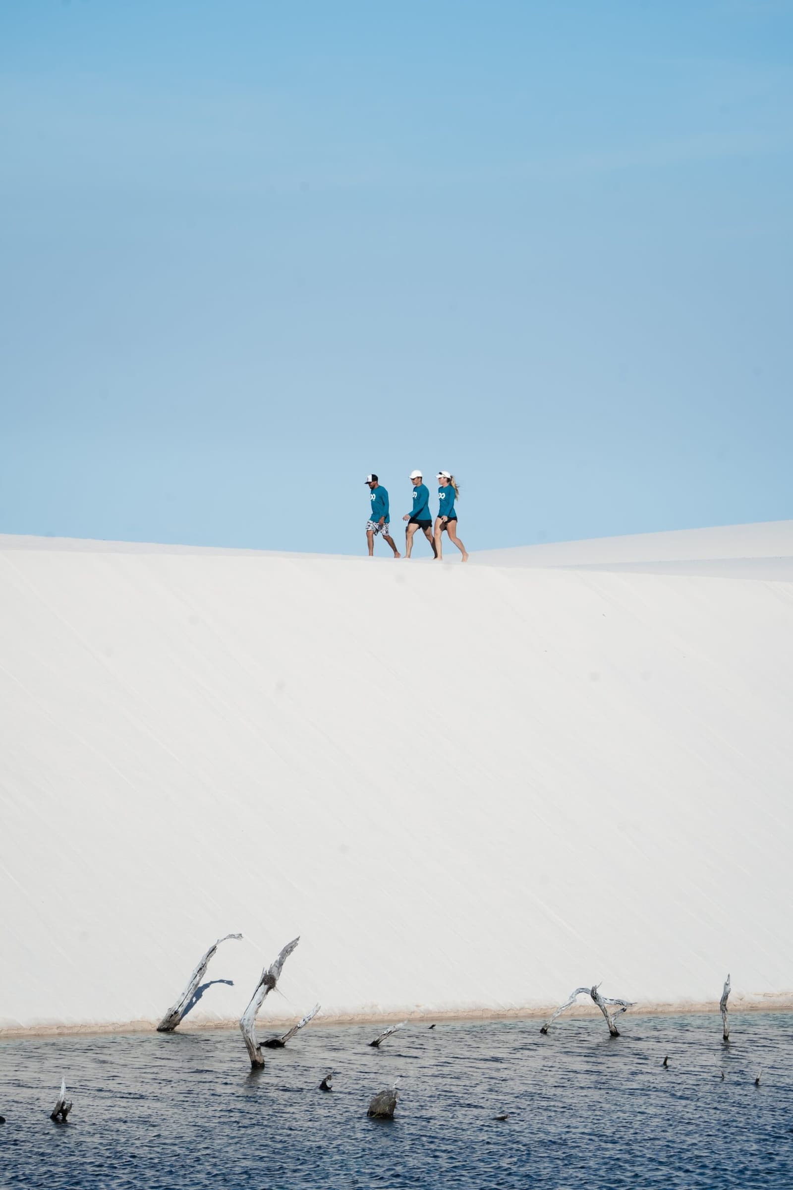 Small figures, enormous dunes, the scale of Lençóis Maranhenses does all the talking. Three trekkers in matching teal shirts walk along the crest of a white sand dune on the Lençóis Maranhenses trek from Atins, with a dark lagoon fringed by bare submerged branches in the foreground and a clear blue sky above