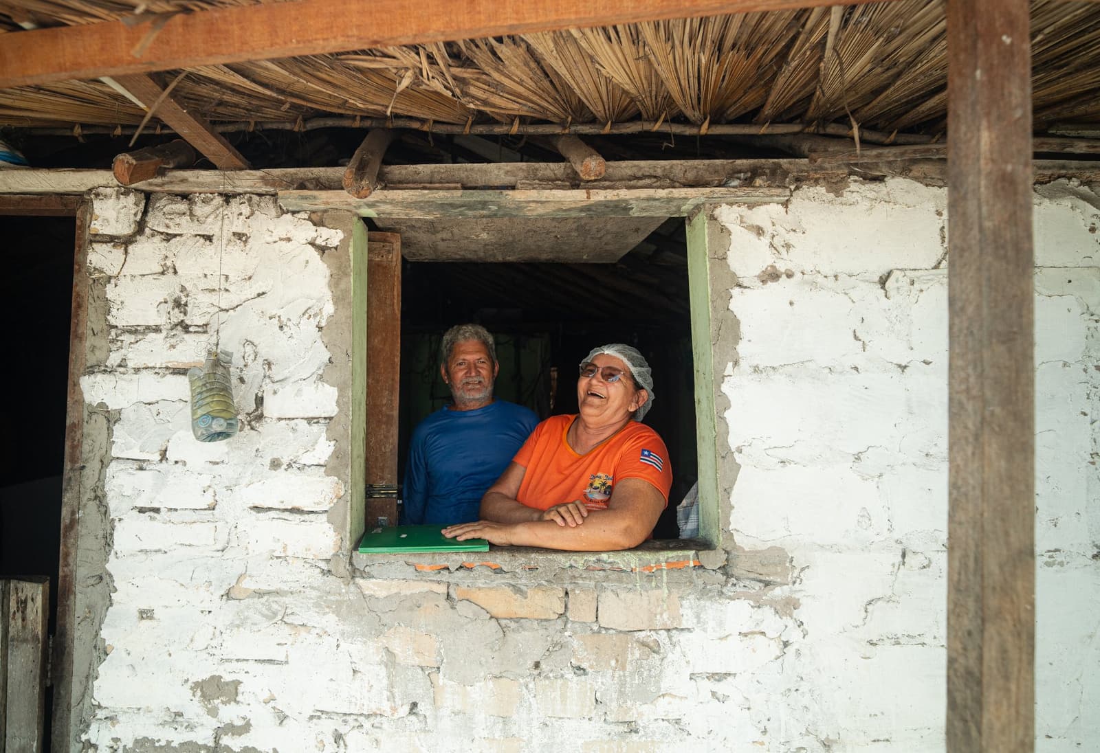 The neighbors always know what's going on before you do. A man in a blue shirt and a woman in an orange shirt and hairnet lean out of a rustic whitewashed brick window with a thatched roof, smiling warmly at the camera during a lencois maranhenses trek homestay.