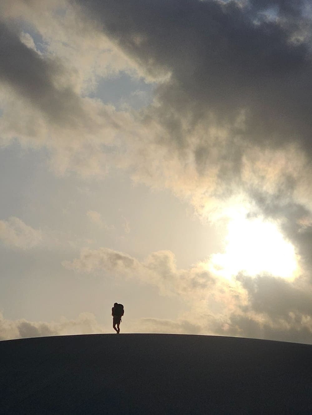 Small figure, very big dune. That feeling is the whole point. Silhouette of a backpacker standing on the curved crest of a white sand dune during the lencois maranhenses trek, under a dramatic cloudy sky with the sun breaking through