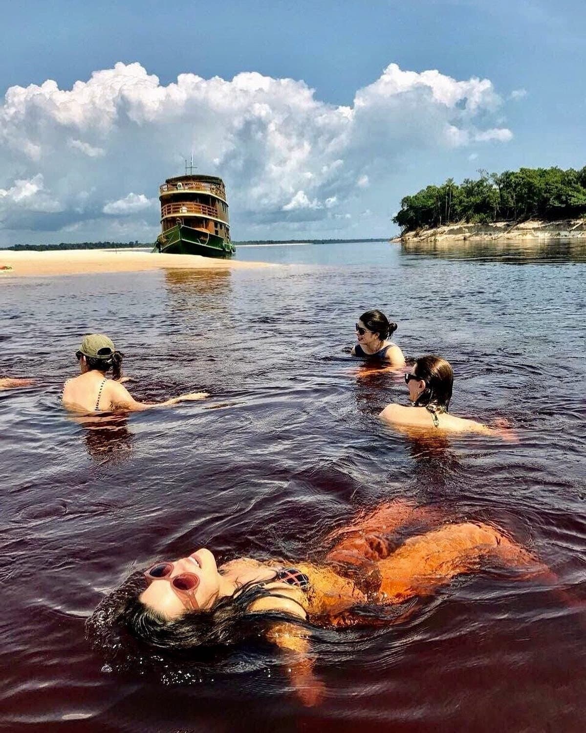 The river is warm, the water is tea-dark, and nobody wants to get back on board. Four travelers swimming in dark tannin-rich black water on an Amazon jungle cruise Brazil, with a vintage multi-deck riverboat anchored beside a sandy beach and lush forest in the background
