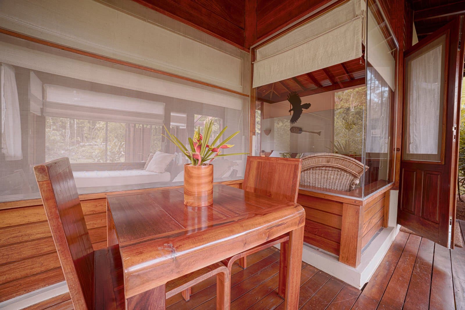 Breakfast at this table after a dawn birding walk is a ritual worth planning a trip around. Photo: Samuel Melim Polished hardwood dining table and chairs on a screened bungalow veranda at a birdwatching in the amazon rainforest lodge, with a tropical flower arrangement in the foreground and a daybed visible through the mosquito netting