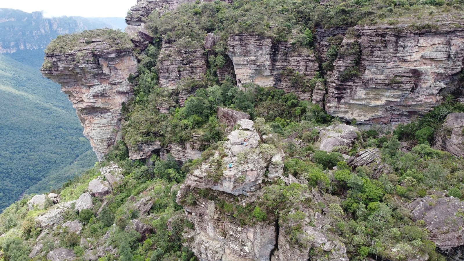 Two dots on a rock ledge, one whole canyon below them. Aerial view of layered sandstone cliffs and green vegetation in the Chapada Diamantina trek, with two tiny hikers visible on an exposed rock outcrop above a deep canyon