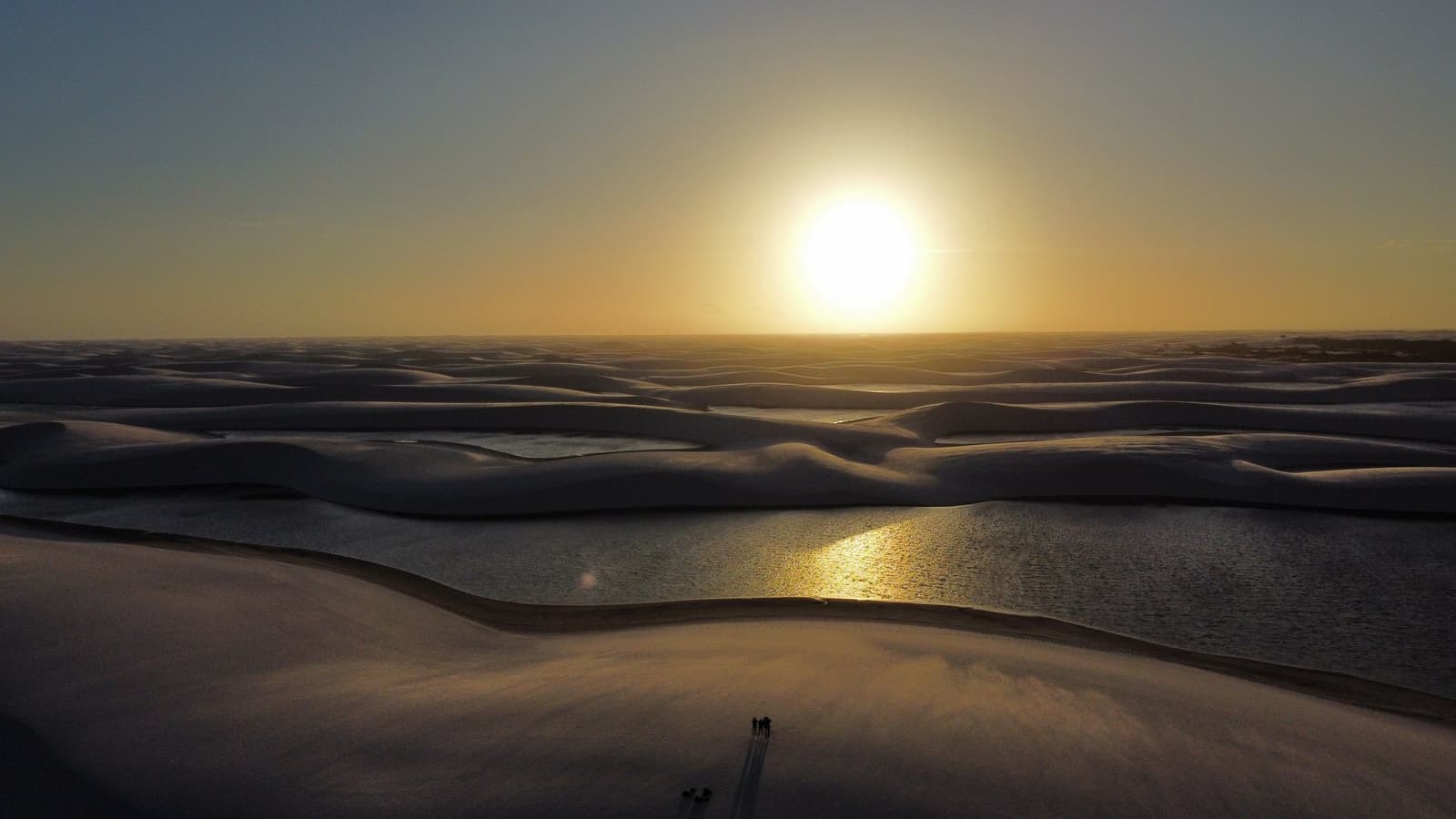 Two people, a sky full of gold, and nowhere else to be. Aerial view of the Lençóis Maranhenses trek landscape at sunset, with white sand dunes casting long shadows across shimmering freshwater lagoons and a small group of hikers visible as tiny silhouettes in the foreground
