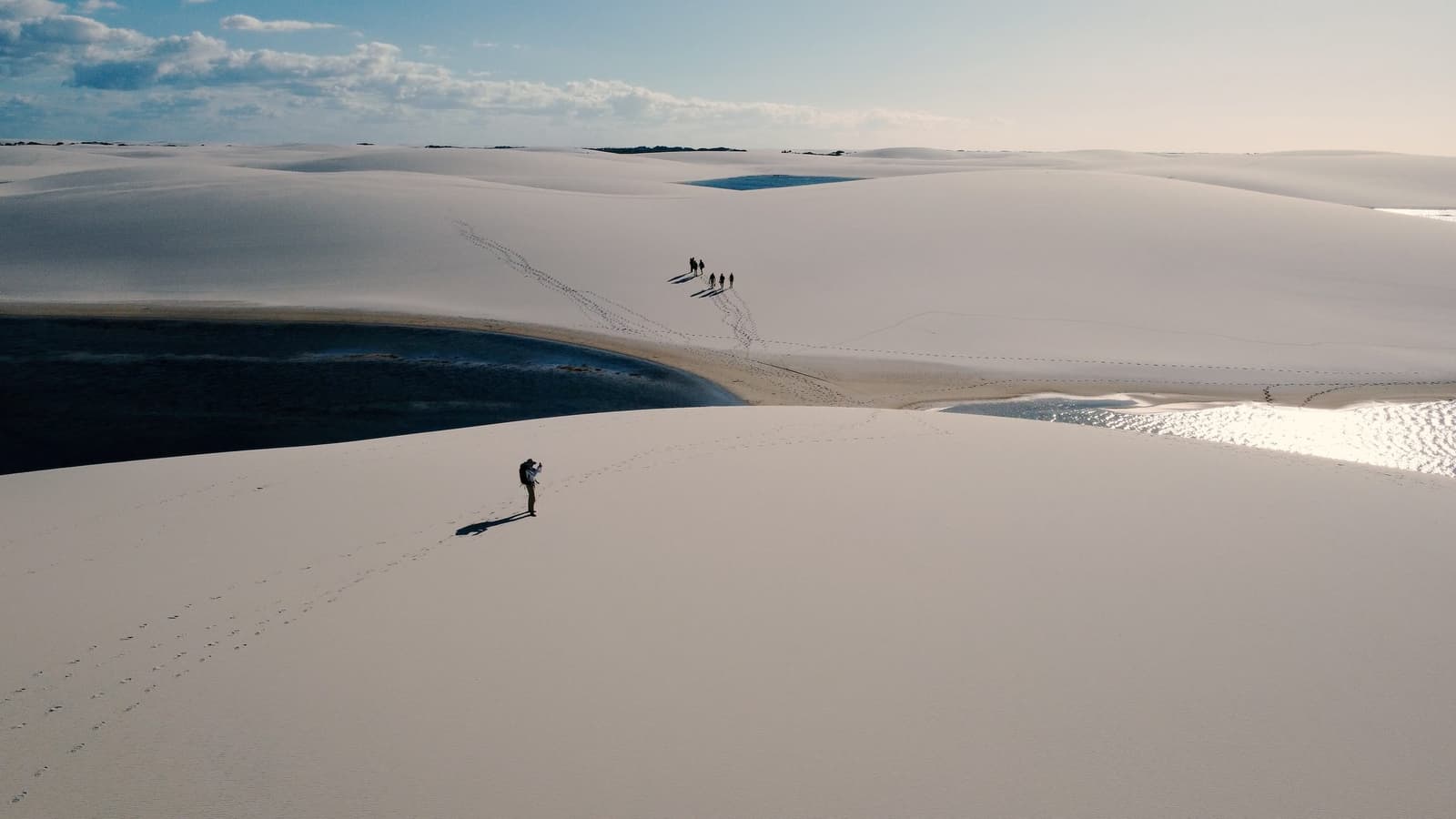 Out here, your footprints are the only map that matters. Aerial view of a lone photographer standing on vast white sand dunes during a Lençóis Maranhenses trekking expedition, with dark-water lagoons reflecting the sky and a small group of hikers visible in the distance
