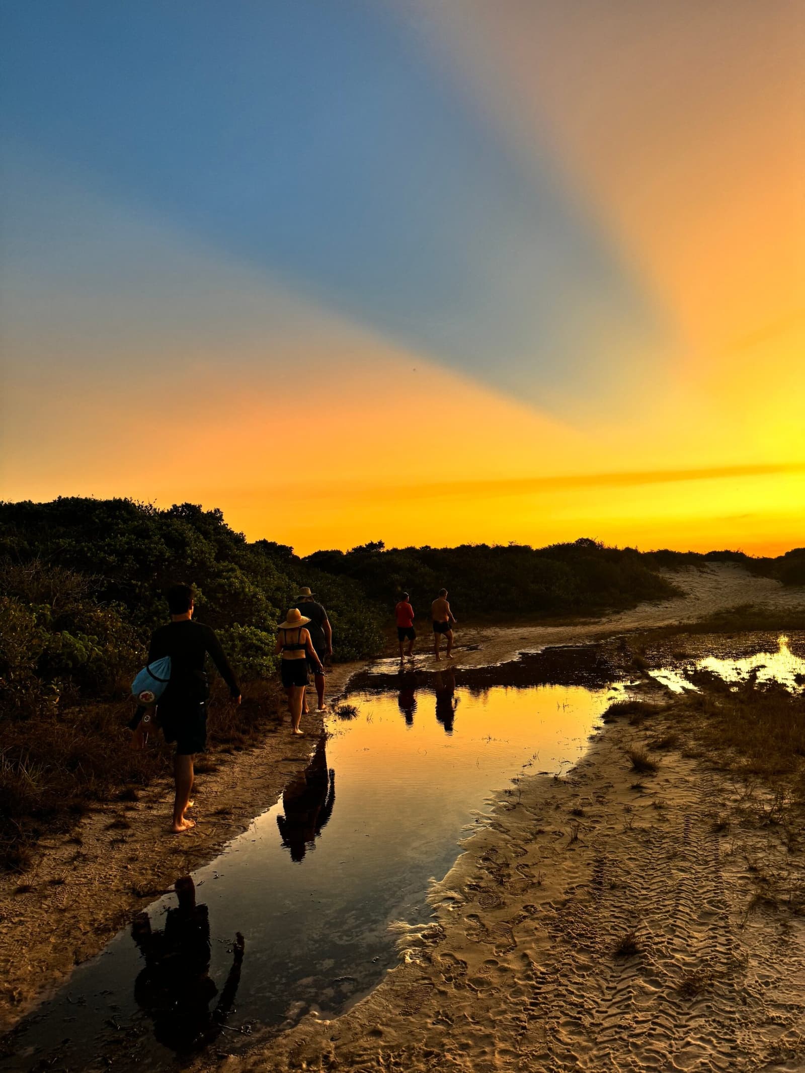 The last light of the day belongs to whoever kept walking. A group of five trekkers walks barefoot along a sandy coastal trail during Lençóis Maranhenses trekking, their silhouettes and the golden sunset sky reflected in a shallow stream cutting through the sand