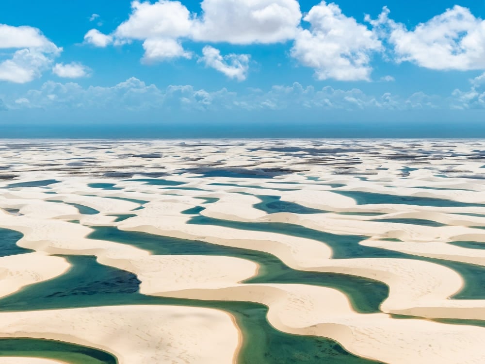 Photo: Julius Dadalti An aerial view of a vast expanse of white sand dunes interspersed with vibrant blue and green seasonal lagoons under a bright blue sky with fluffy white clouds in Lençóis Maranhenses National Park, Brazil.