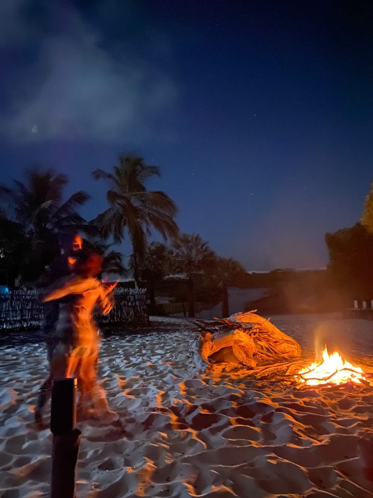 Sand still warm, fire lit, the night just getting started. A person in motion beside a crackling bonfire on a white-sand beach at night, palm trees silhouetted against a deep blue sky, during the Lençóis Maranhenses trek