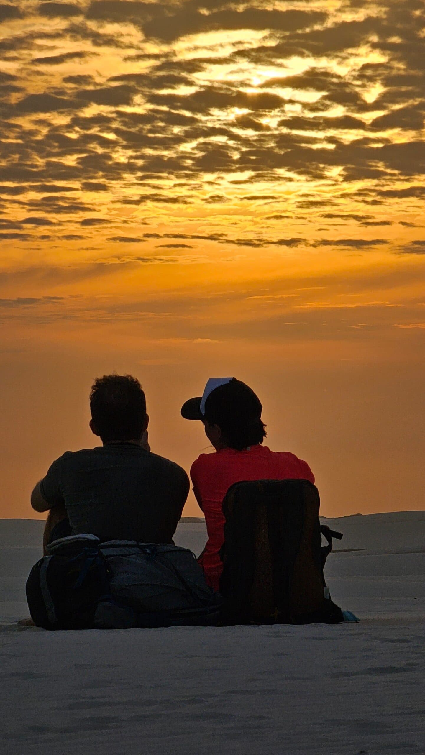 No agenda, no signal, just that sky doing its thing. Two travelers sit side by side on white sand dunes during a Lençóis Maranhenses trekking sunset, silhouetted against a golden-orange sky filled with scattered clouds