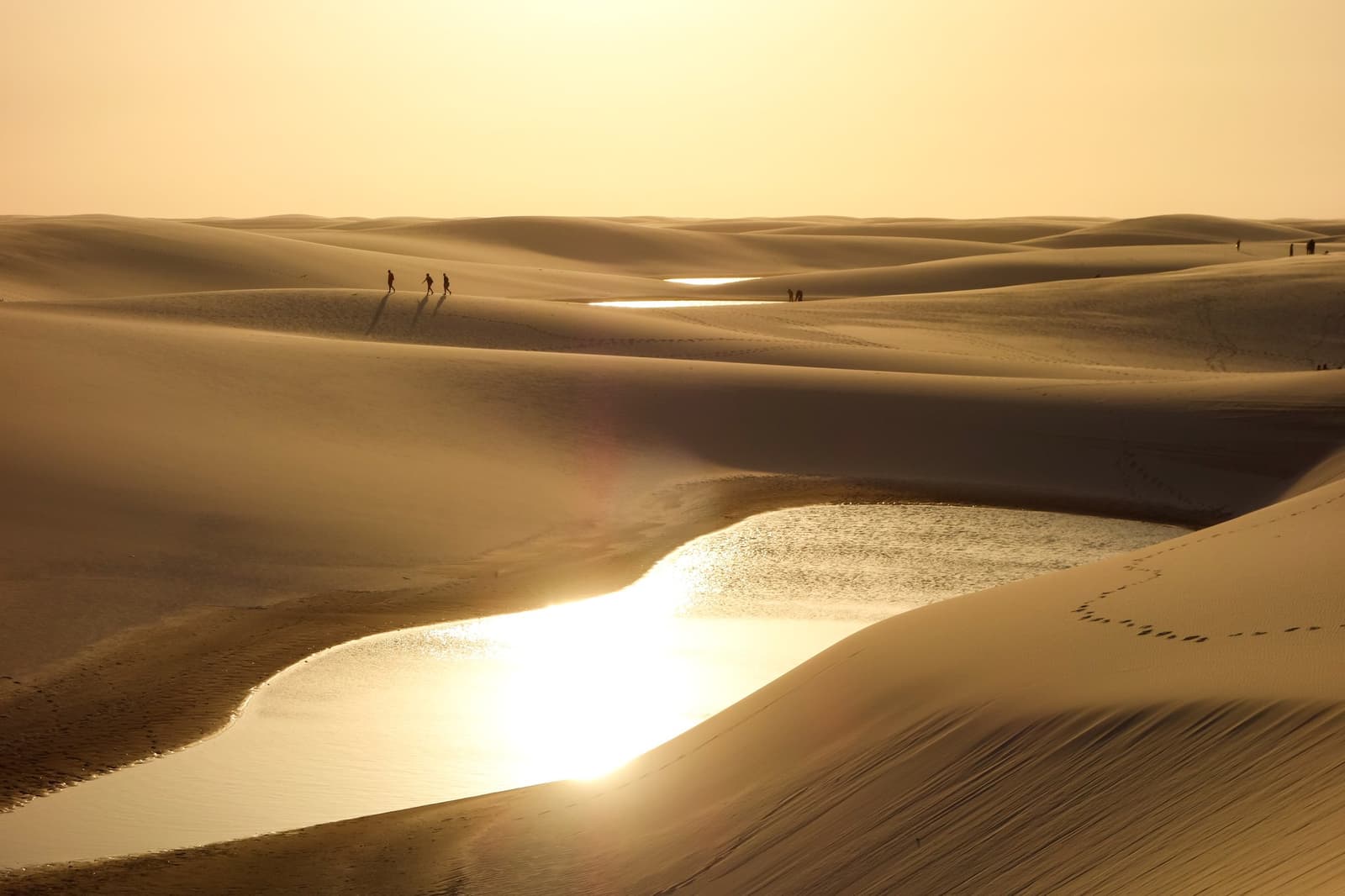 A desert that drinks. The lagoons appear and the whole place shifts. Three silhouetted hikers walk the crest of a golden dune during a Lençóis Maranhenses hike, with shallow rain-filled lagoons glittering in the low evening light across the vast dune field