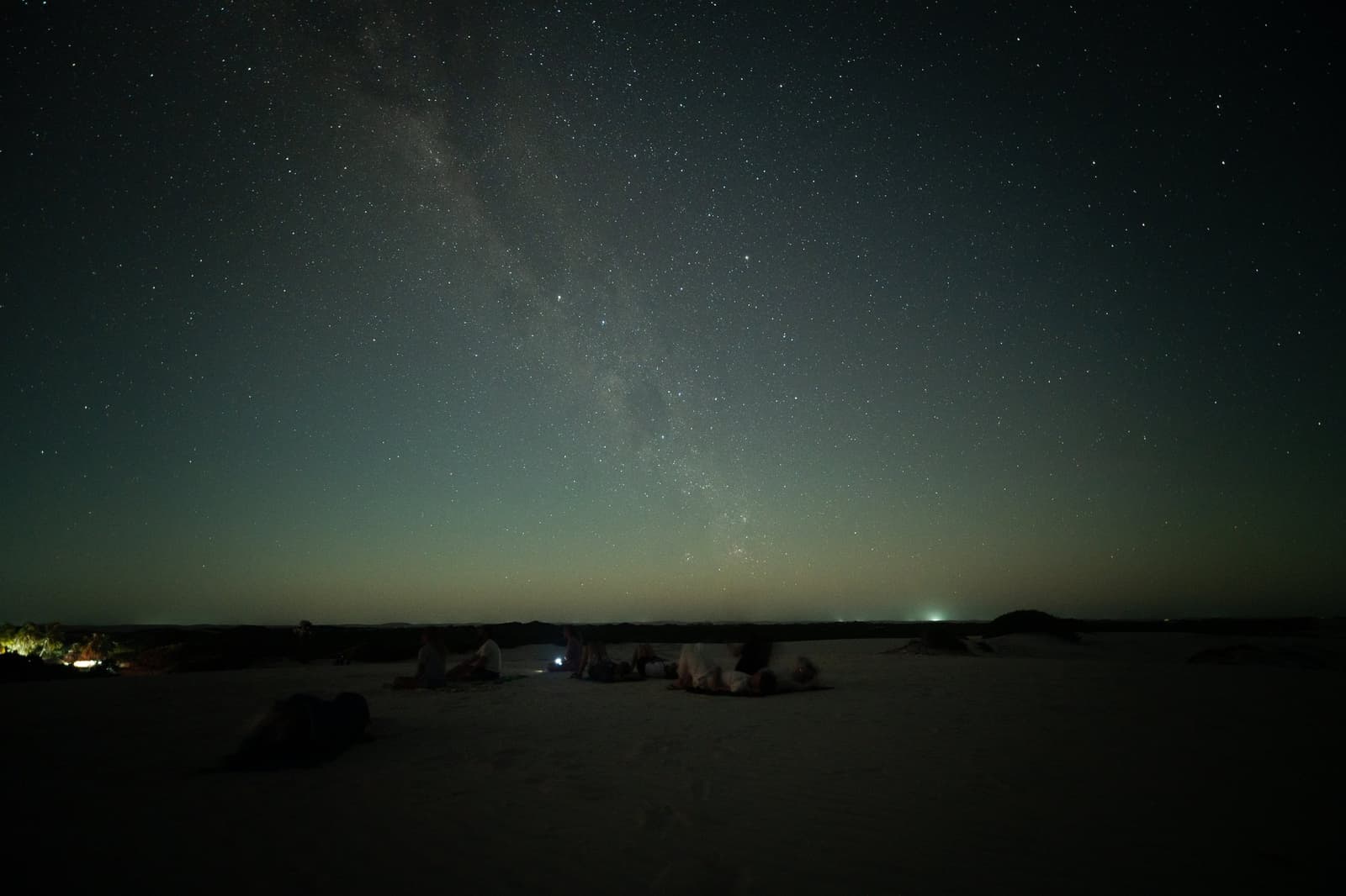 No light pollution, no roof, just the whole galaxy showing off. A small group of travelers lying on white sand dunes watching the Milky Way arc across a dark, star-filled sky on a lencois maranhenses trek, with a faint glow on the horizon