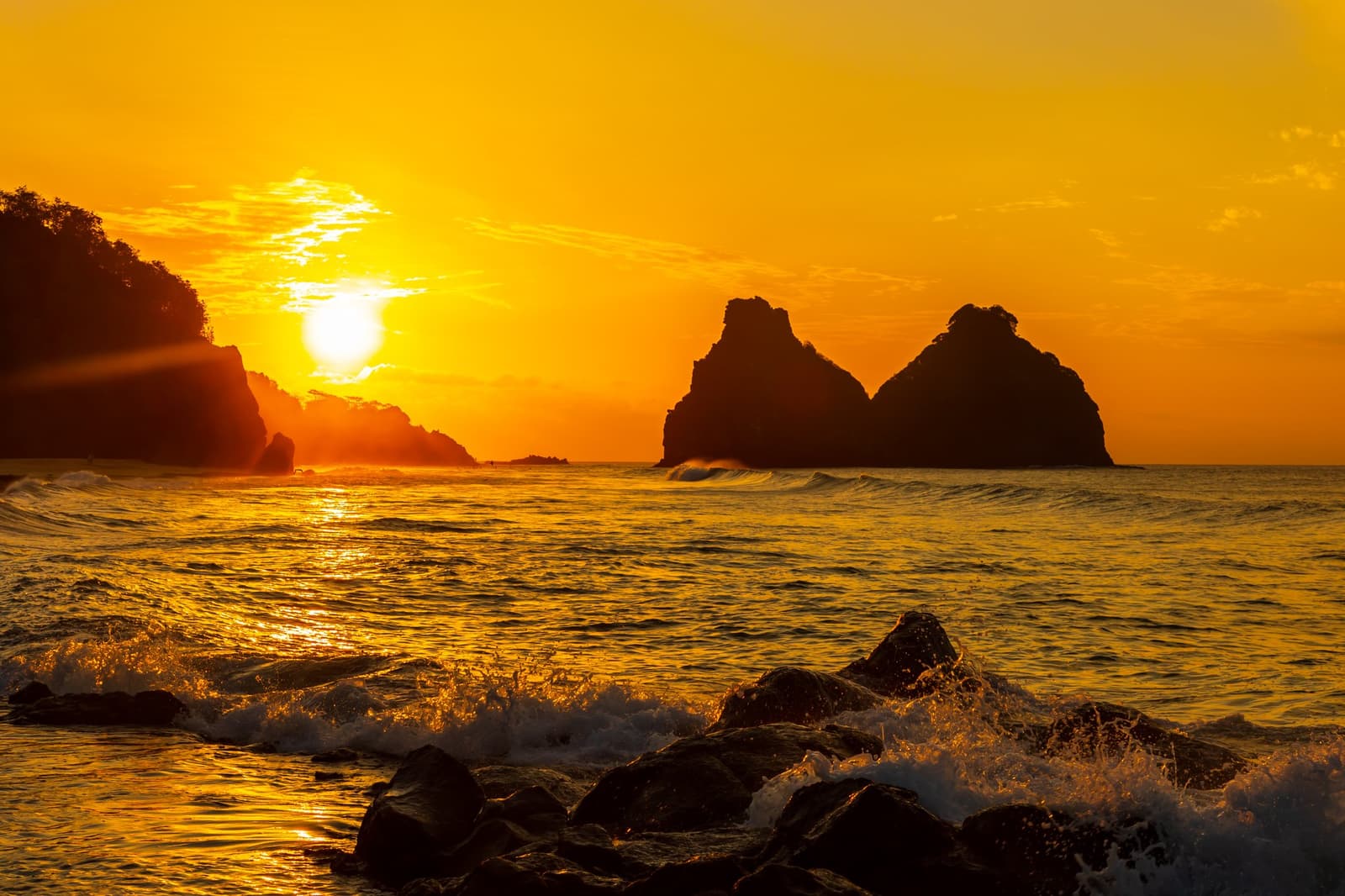 The kind of ending to a dive day that makes you forget to check your phone. Fiery golden sunset over silhouetted rocky sea islets off Fernando de Noronha, waves crashing over foreground boulders, part of a 2-Day Diving Package in Fernando de Noronha
