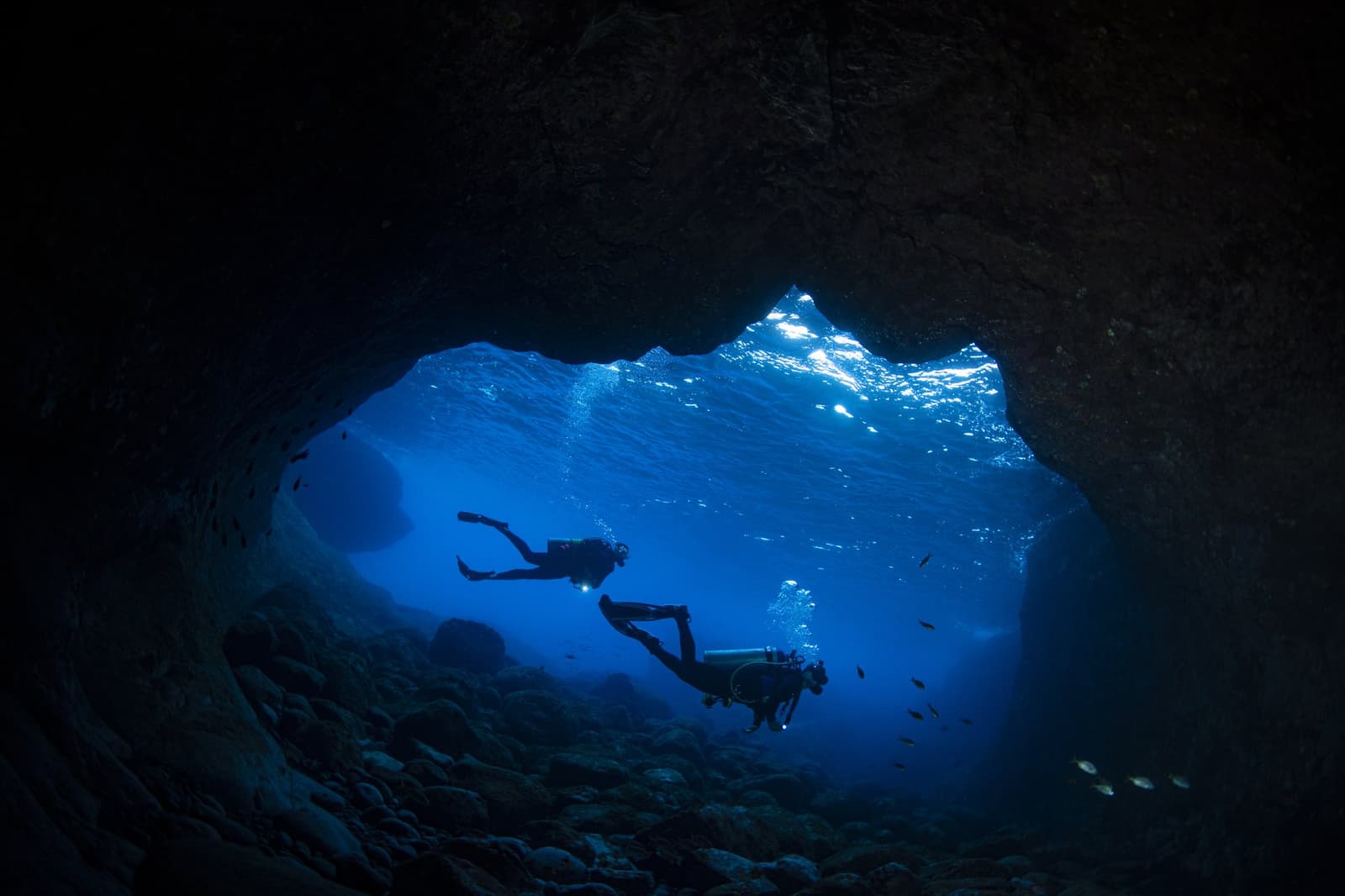 The cave does the dramatic work, the divers just get to live it. Two scuba divers silhouetted against brilliant blue water inside a rocky underwater cave entrance, part of the 2-Day Diving Package in Fernando de Noronha