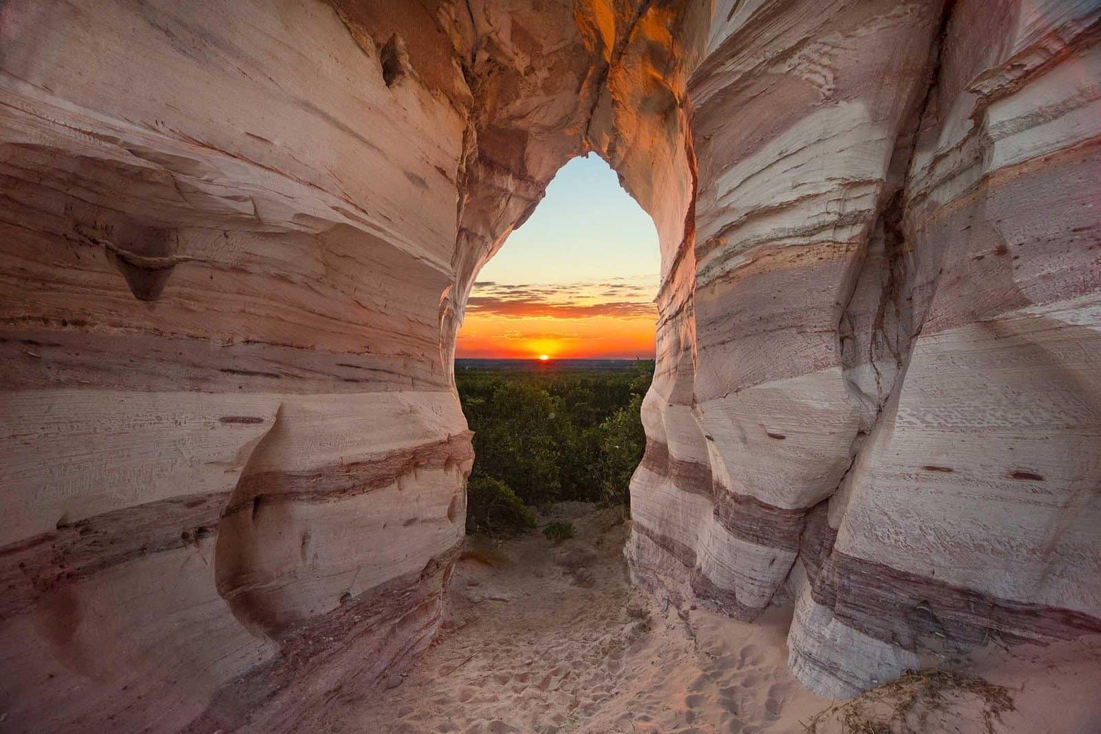 Fiery dusk pulled through the eye of ancient stone. Warm orange sunset framed by the natural stone arch of Pedra Furada, a striped sandstone formation in Jalapão, with green treetops visible below on the 4-day Jalapão tour