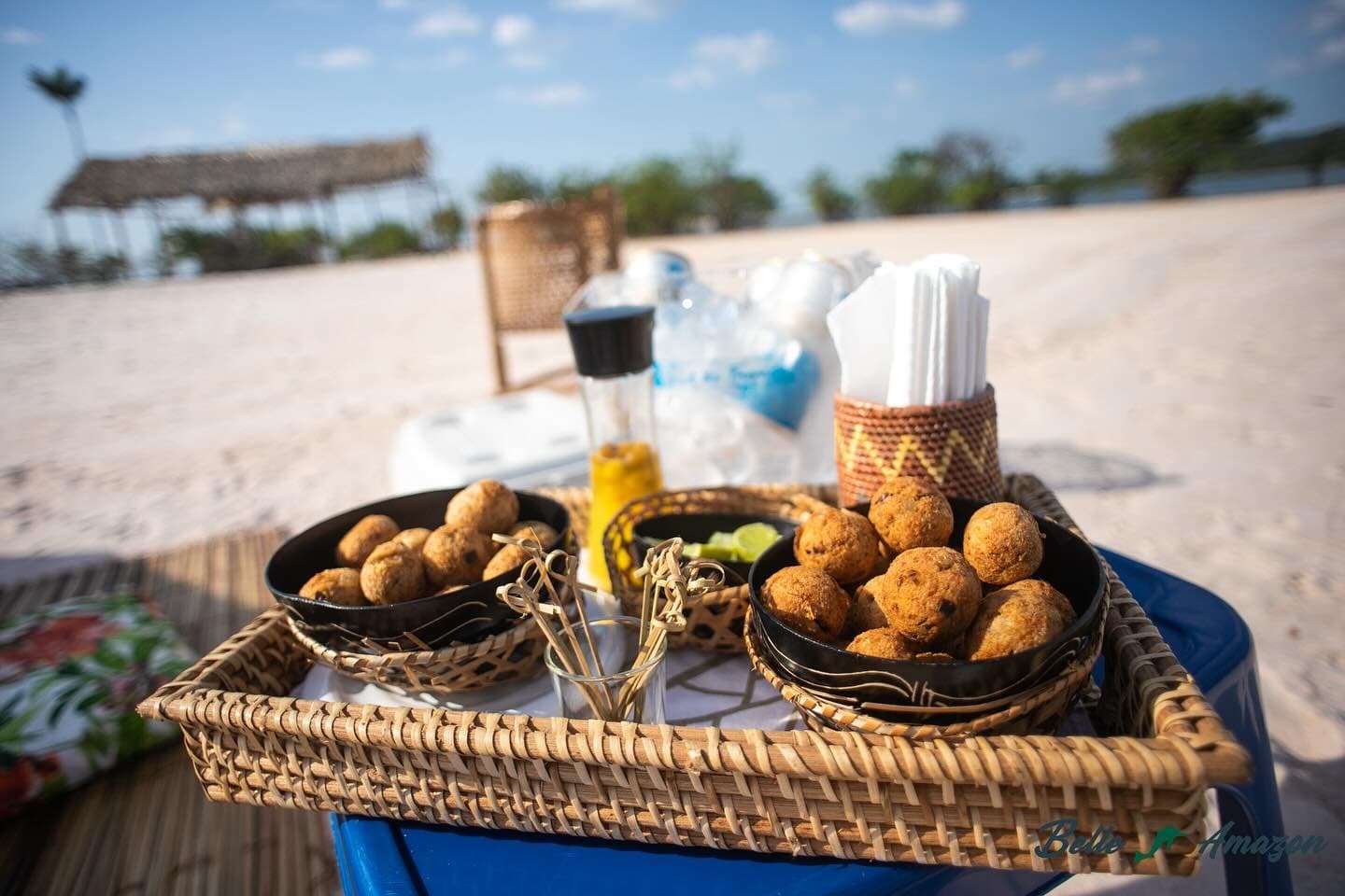 Snack hour on the sandbank: the Amazon has its own ideas about fine dining. A woven rattan tray holding two black bowls of golden fried snack balls served on a sandy riverbank during an Amazon cruise from Alter do Chão, with bamboo skewers, lime wedges and condiments alongside