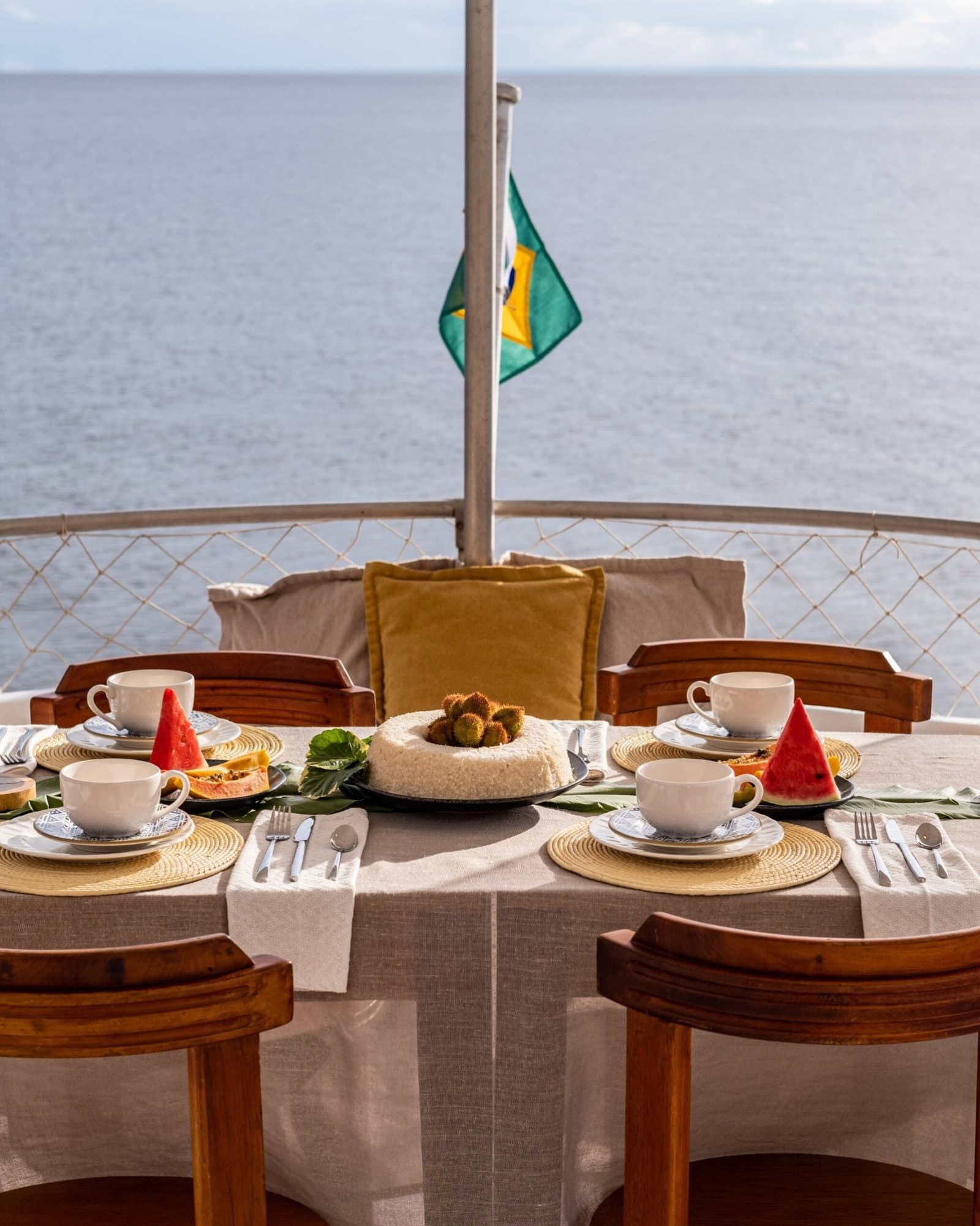 Breakfast with a river view and nowhere else to be. A set breakfast table on the open deck of an Amazon cruise from Alter do Chão, with fresh watermelon, papaya, white coffee cups, and a centrepiece cake, a Brazilian flag flying over calm open water in the background