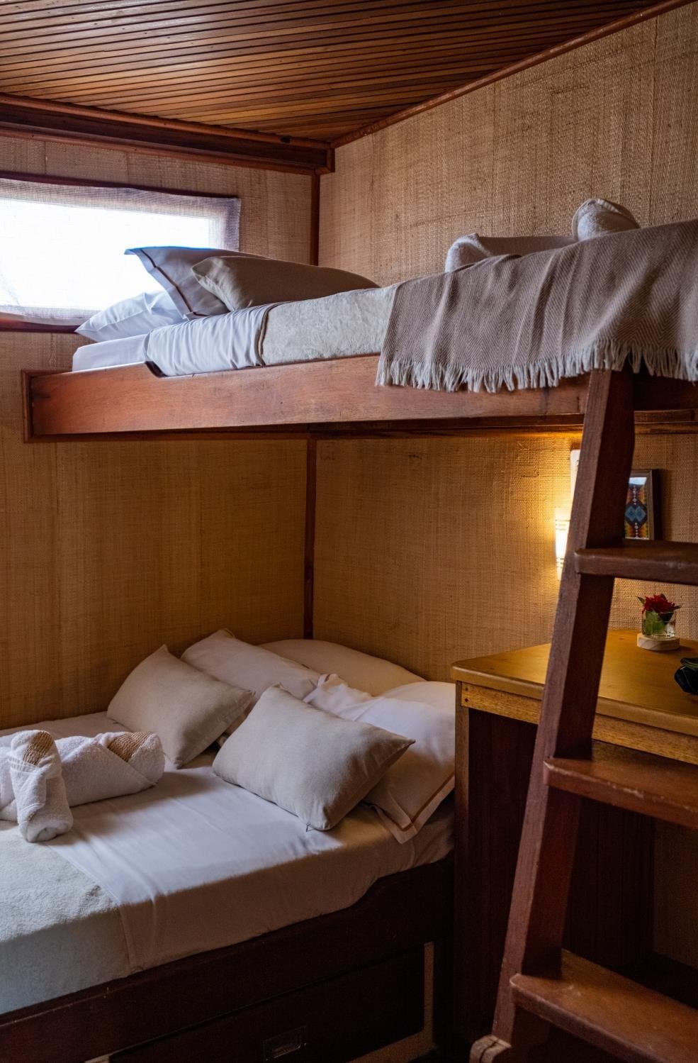 Compact, yes, but after a day on the river, this feels just right. Cosy wooden bunk cabin aboard an Amazon cruise from Alter do Chão, with white linen, cushioned pillows, a fringed throw on the upper bunk, warm bedside lighting, and a timber ladder