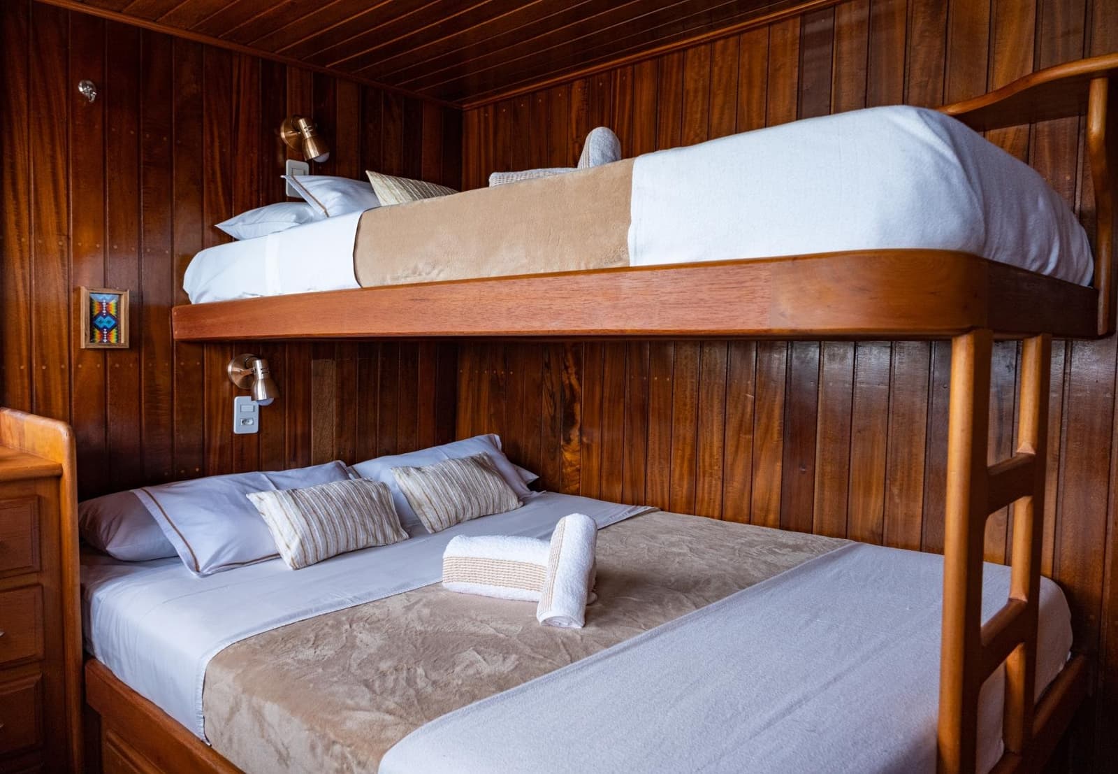 Compact, warm, and surprisingly easy to sleep in, the river does the rest. A polished timber bunk cabin aboard an Amazon cruise from Alter do Chão, featuring a double lower berth dressed in crisp white and taupe linen, an upper bunk, brass reading lamps, and warm dark-wood panelling throughout