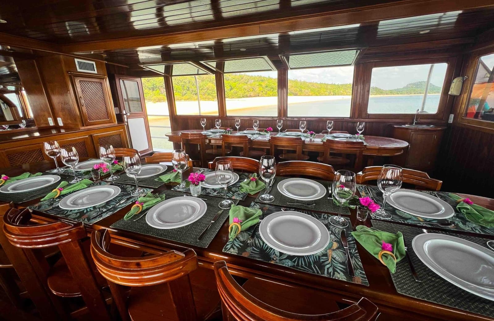Dinner with a front-row view of the river. Not bad for a Tuesday. Elegantly set dining tables with wine glasses, green napkins and pink floral accents inside a polished wood-panelled salon on an Amazon cruise from Alter do Chão, with a sandy river beach and forested hills visible through panoramic windows