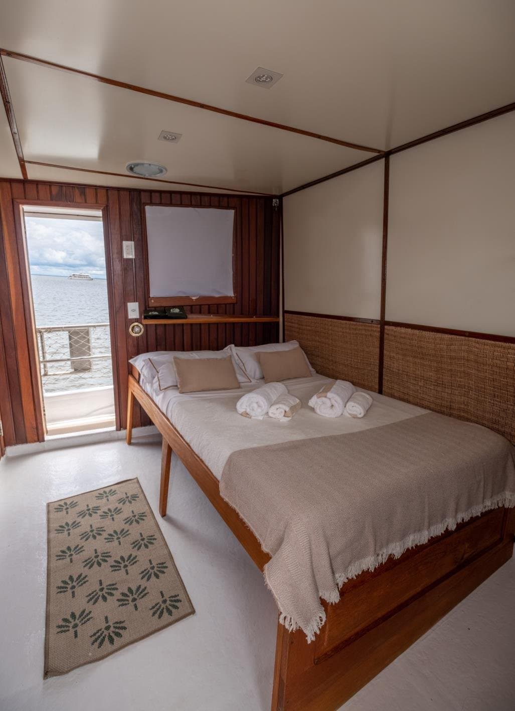 Your own slice of the river, right outside the cabin door. A neatly made double bed with rolled white towels on a woven-headboard cabin aboard an Amazon cruise from Alter do Chão, with an open door revealing a water view and cloudy sky beyond