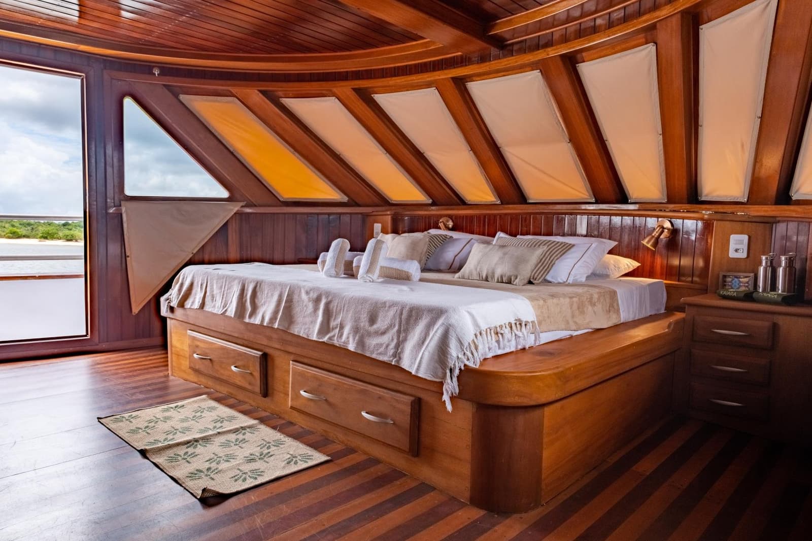 Your floating home on the Amazon sleeps as well as it travels. Polished hardwood master cabin aboard a luxury Amazon cruise from Alter do Chão, featuring a built-in double bed with storage drawers, canvas skylights, and an open side door revealing a riverside landscape