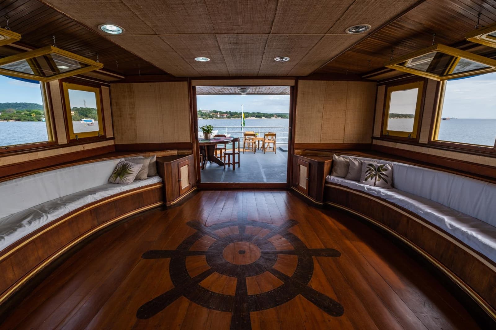 The ship's wheel on the floor is just showing off, honestly. Polished hardwood salon aboard an Amazon cruise from Alter do Chão, with curved bench seating, a ship's-wheel floor inlay and open doors leading to a rear deck overlooking a broad river
