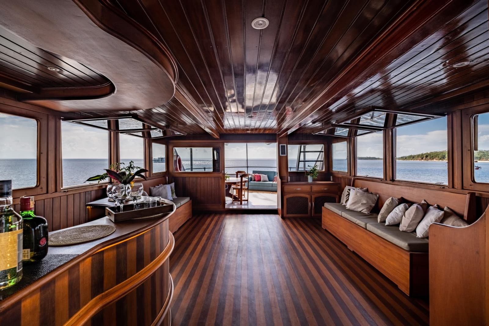 Your living room for the week, with considerably better views. Polished hardwood salon aboard a luxury Amazon cruise from Alter do Chão, with built-in cushioned seating, a bar tray set with glasses, striped teak flooring, and panoramic river views through wraparound windows