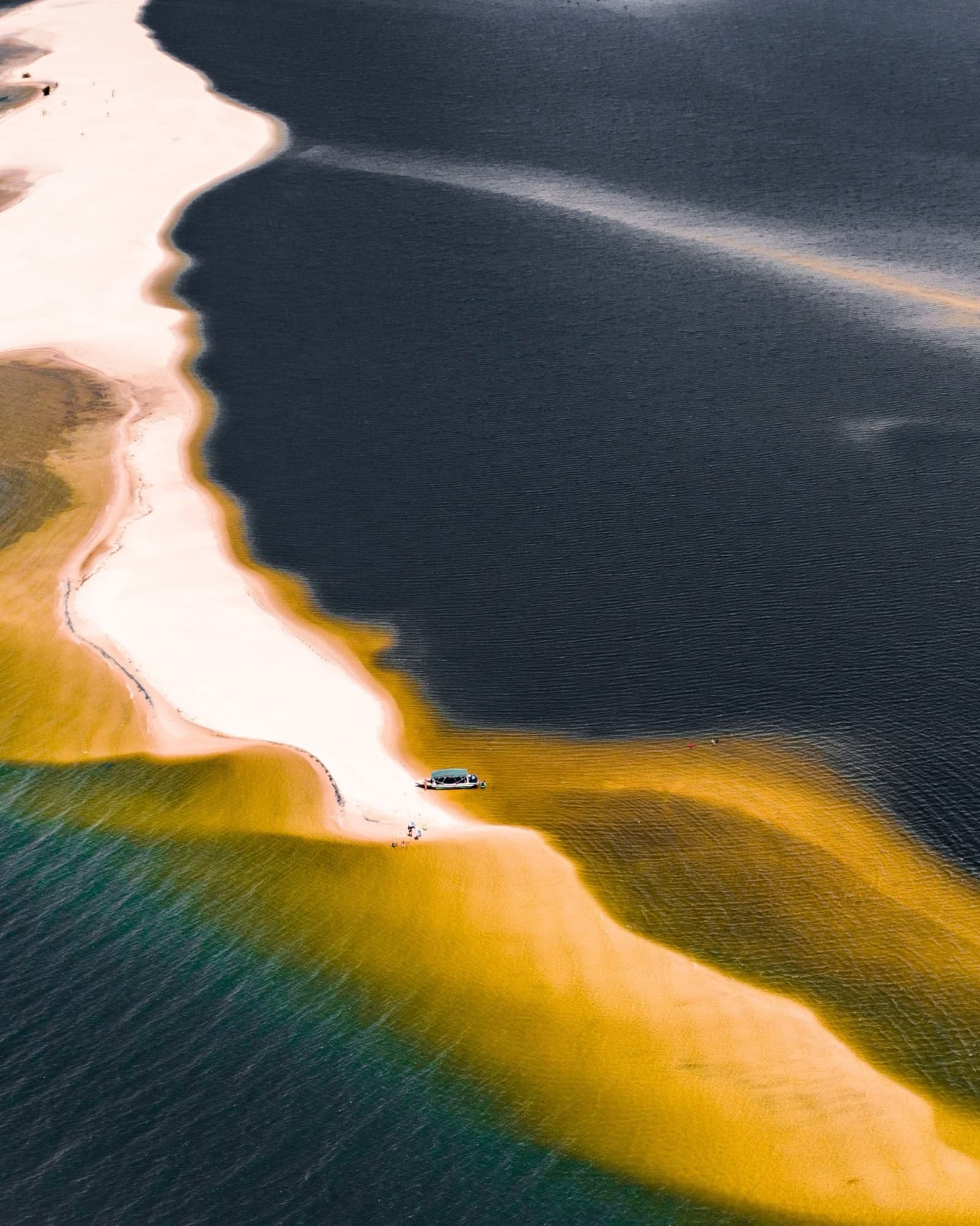 Three colors of water, one sliver of sand, and a boat that got there first. Aerial view of a small boat moored on a winding white sandbar where teal, gold and dark river waters meet during an Amazon cruise from Alter do Chão