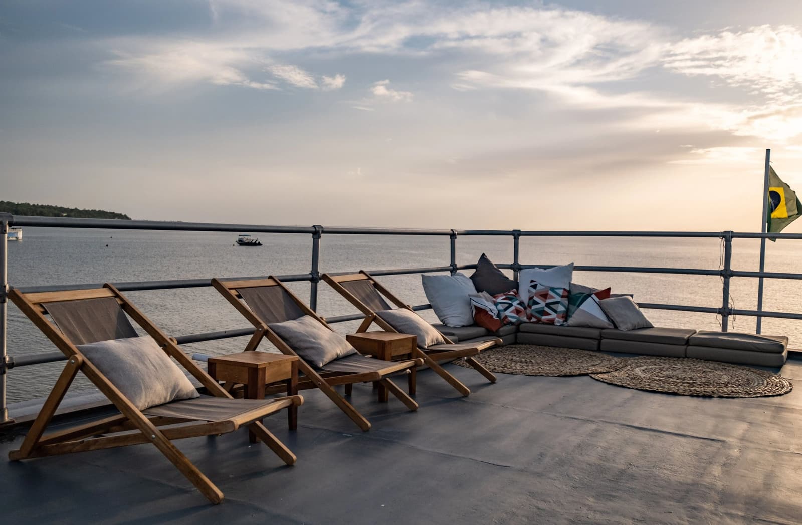 The best seat on the river, and it's yours all evening. Teak deck chairs and a cushioned lounge corner on the upper deck of an Amazon cruise from Alter do Chão, with a broad river stretching to the horizon under a soft evening sky and the Brazilian flag flying at the stern