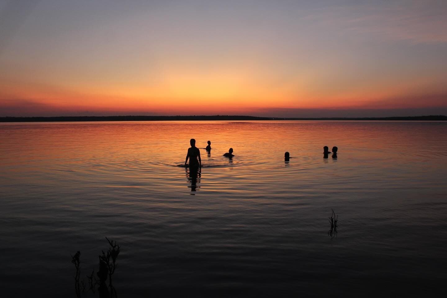 The river keeps the day going a little longer, if you let it. Silhouettes of travellers swimming in a vast, glassy river at dusk on an Amazon cruise from Alter do Chão, with a fiery orange and red sunset reflected across the water