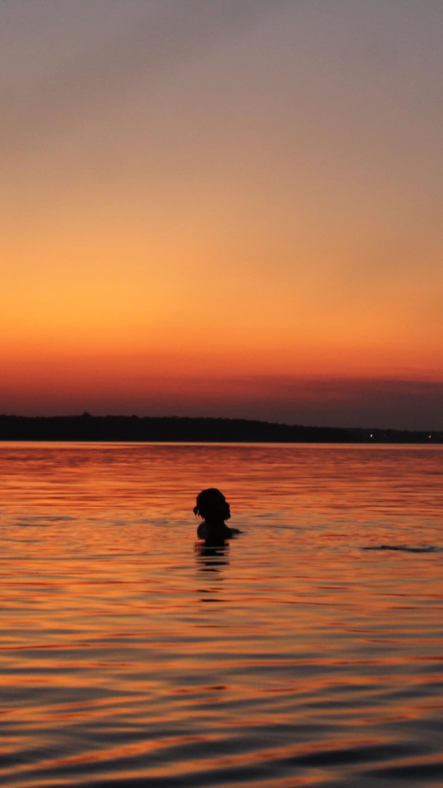 Some sunsets are best felt from the inside out. A person swimming in a vast river at dusk, silhouetted against a deep orange sunset sky on an Amazon cruise from Alter do Chão