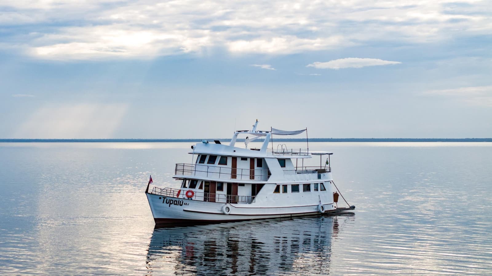 Your floating base camp, ready and waiting on the river. The Tupaiu vessel, part of the Amazon cruise from Alter do Chão fleet, moored on a glassy broad river under a soft overcast sky