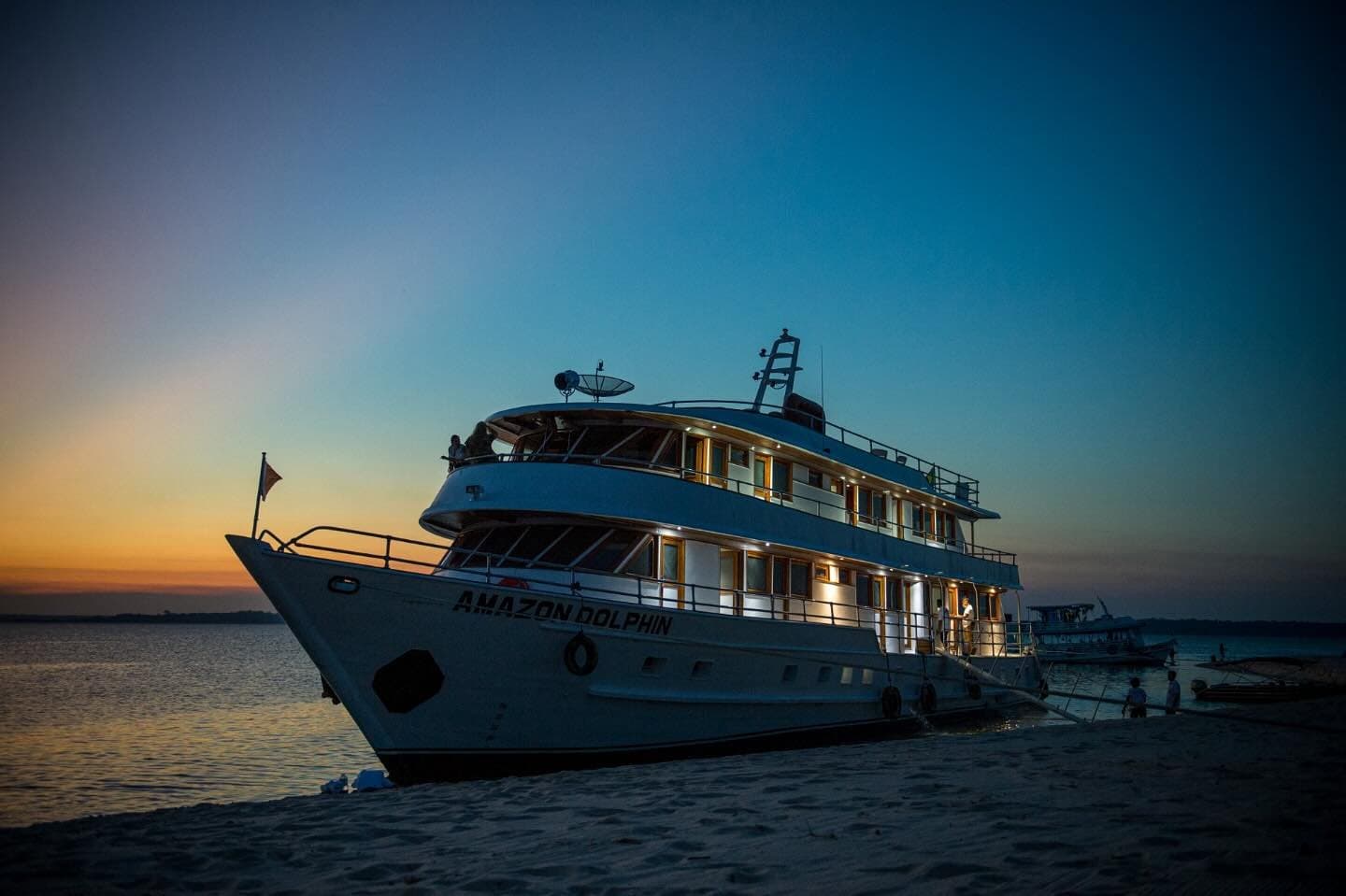 Your floating base camp, ready and lit before the river goes dark. The Amazon Dolphin cruise vessel moored on a river sandbar at dusk, warm cabin lights glowing against a deep blue and orange sunset sky on an Amazon cruise from Alter do Chão