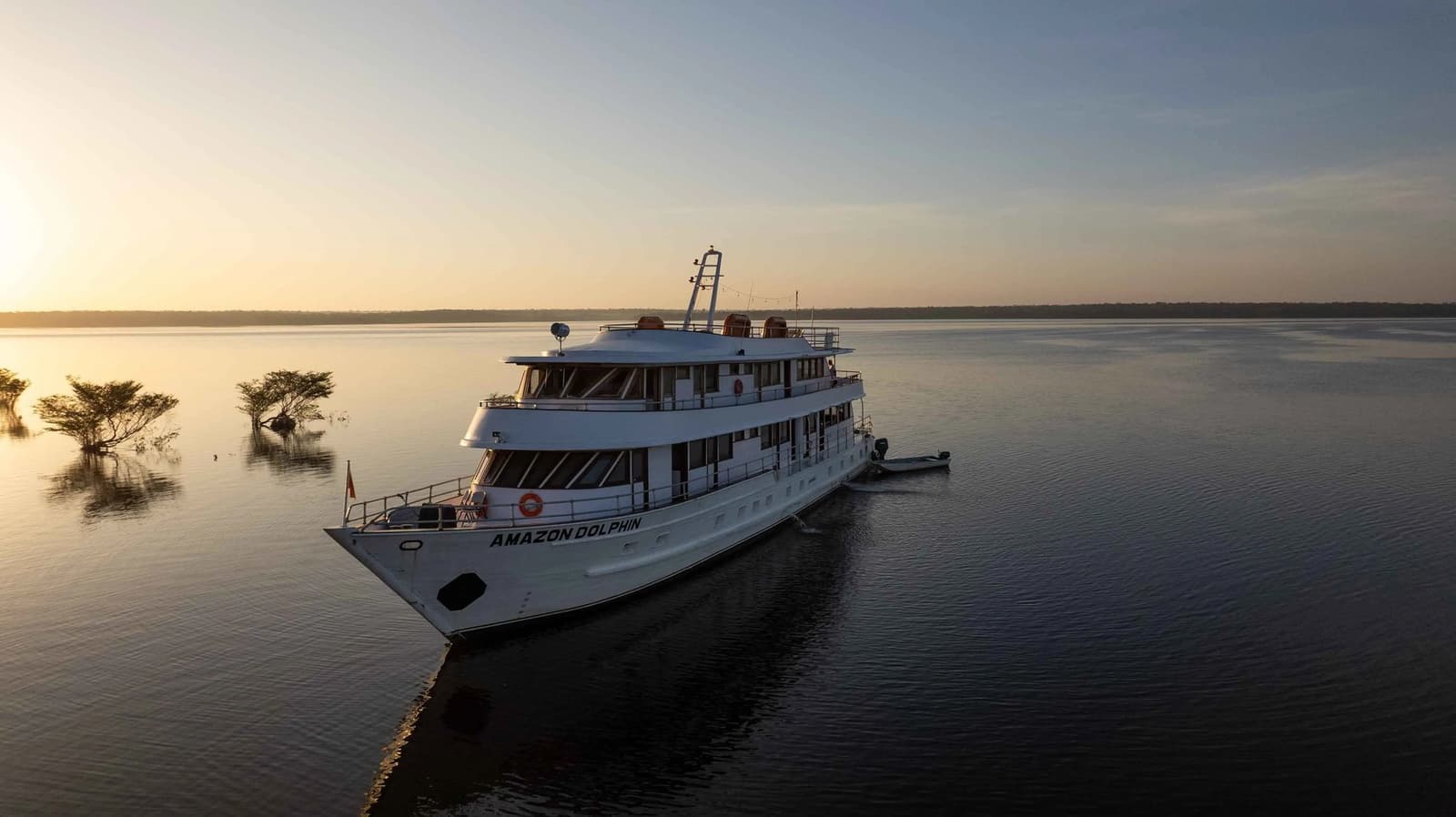 Your home on the river, right where the sky catches fire. The Amazon Dolphin vessel anchored on a glassy river at golden-hour sunset, part of the Amazon cruise from Alter do Chão fleet, with flooded shoreline trees silhouetted against a warm horizon