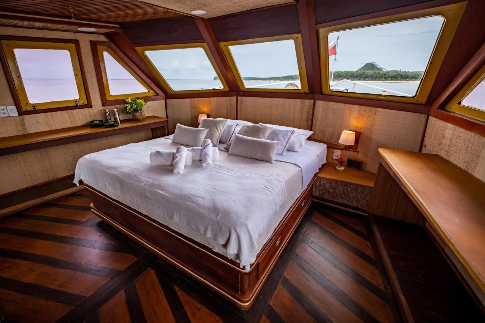 Your floating bedroom comes with a view that changes every morning. Spacious master cabin aboard an Amazon cruise from Alter do Chão, with a king-size bed dressed in crisp white linen, towel-folded animals, warm timber panelling, and panoramic windows overlooking a forested riverbank