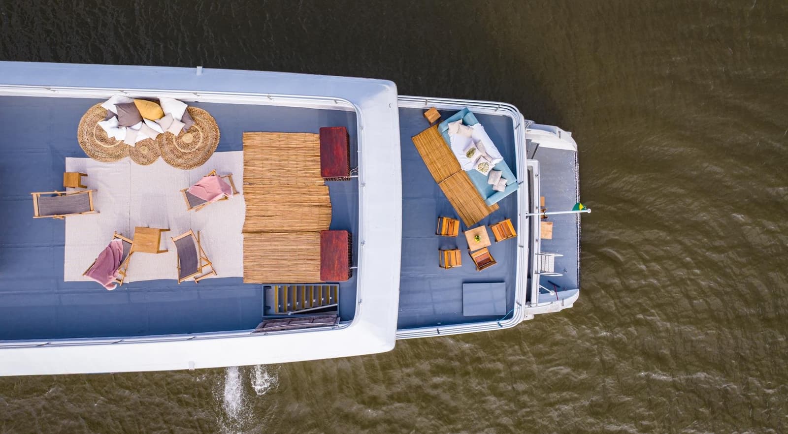 Your living room for the next few days, just happens to float. Aerial view of a luxury Amazon cruise from Alter do Chão showing two open sundecks furnished with wooden loungers, rattan rugs, thatched shade structures and cushioned seating, cruising across a wide brown river