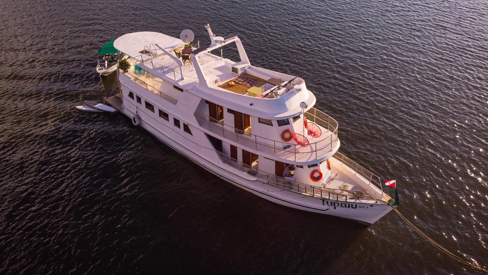 Your floating base camp, ready for whatever the river has next. Aerial view of the white multi-deck Tupaiu river vessel on a broad dark river, with open sun decks, teak lounging areas and a smaller tender tied alongside — the Amazon cruise from Alter do Chão in full.