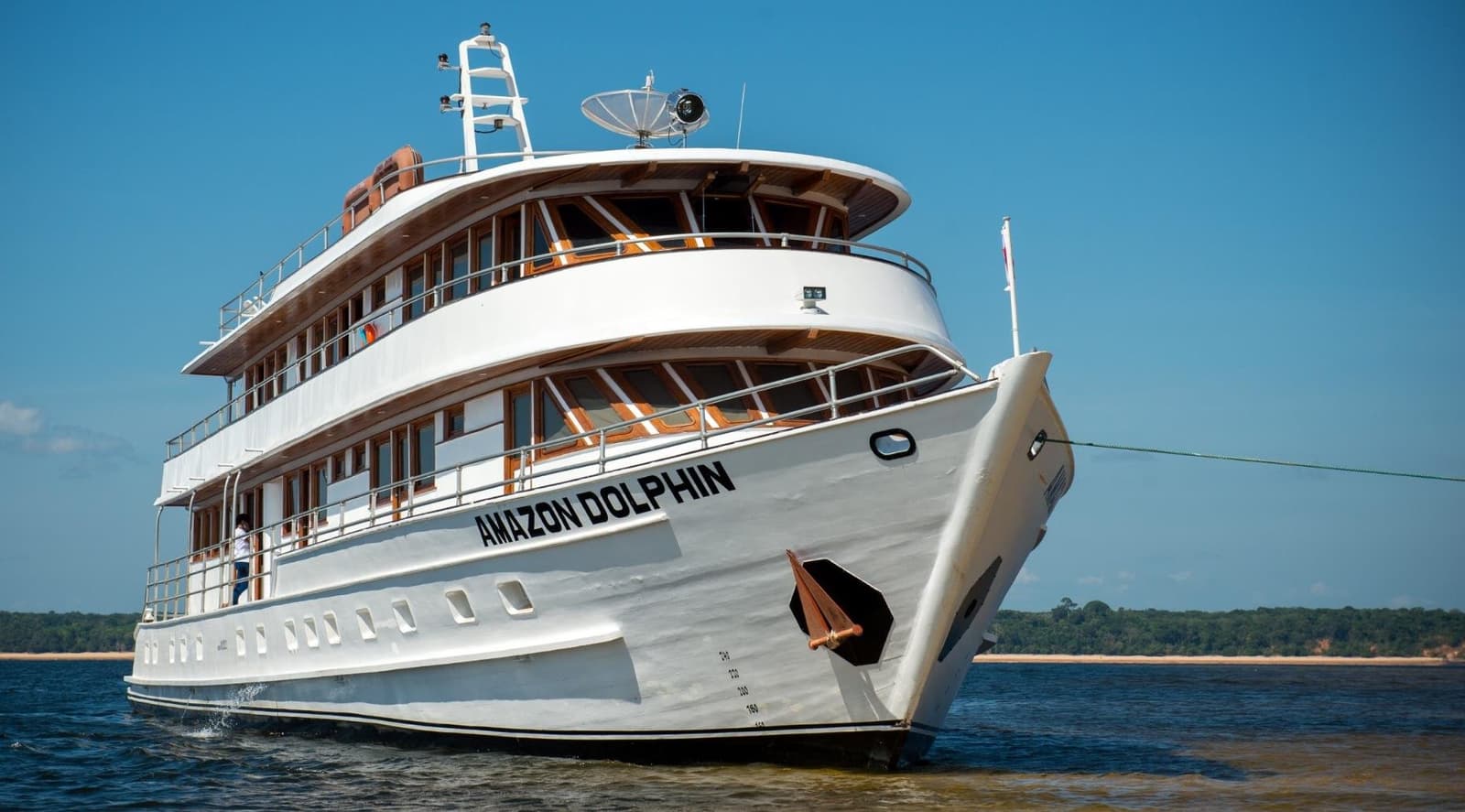 Your floating base camp, named after the river's most charismatic resident. The Amazon Dolphin multi-deck river cruise vessel underway on a wide river channel, with warm teak woodwork and white hull reflecting the bright blue sky — part of the Amazon cruise from Alter do Chão experience