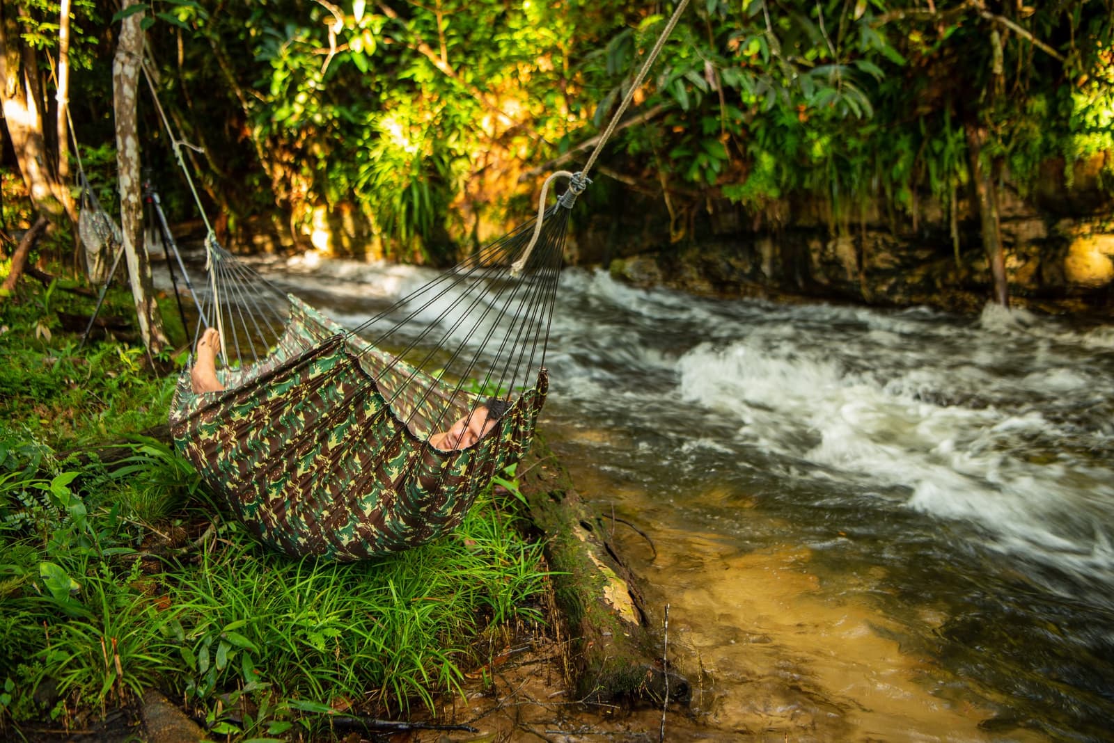 The river does the talking; you just lie back and listen. A traveler resting in a camouflage hammock strung beside a rushing jungle stream, surrounded by lush tropical foliage on an Amazon kayak tour in Brazil