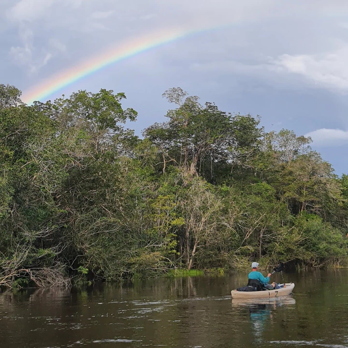 Not every paddle stroke comes with a rainbow, but some do. A solo kayaker paddles toward dense rainforest trees along a dark-water river on an Amazon kayak tour in Brazil, with a vivid rainbow arching across the stormy sky above