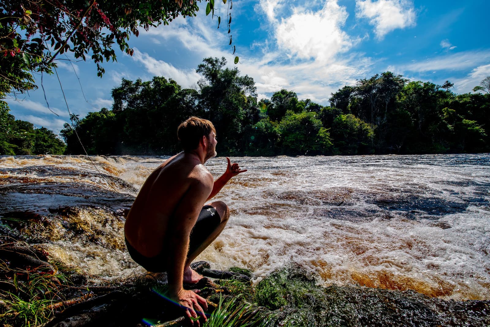 Sizing up the rapids before the river makes the decision for you. A traveler crouches at the rocky edge of churning brown rapids on an Amazon kayak tour in Brazil, surrounded by dense rainforest under a bright blue sky