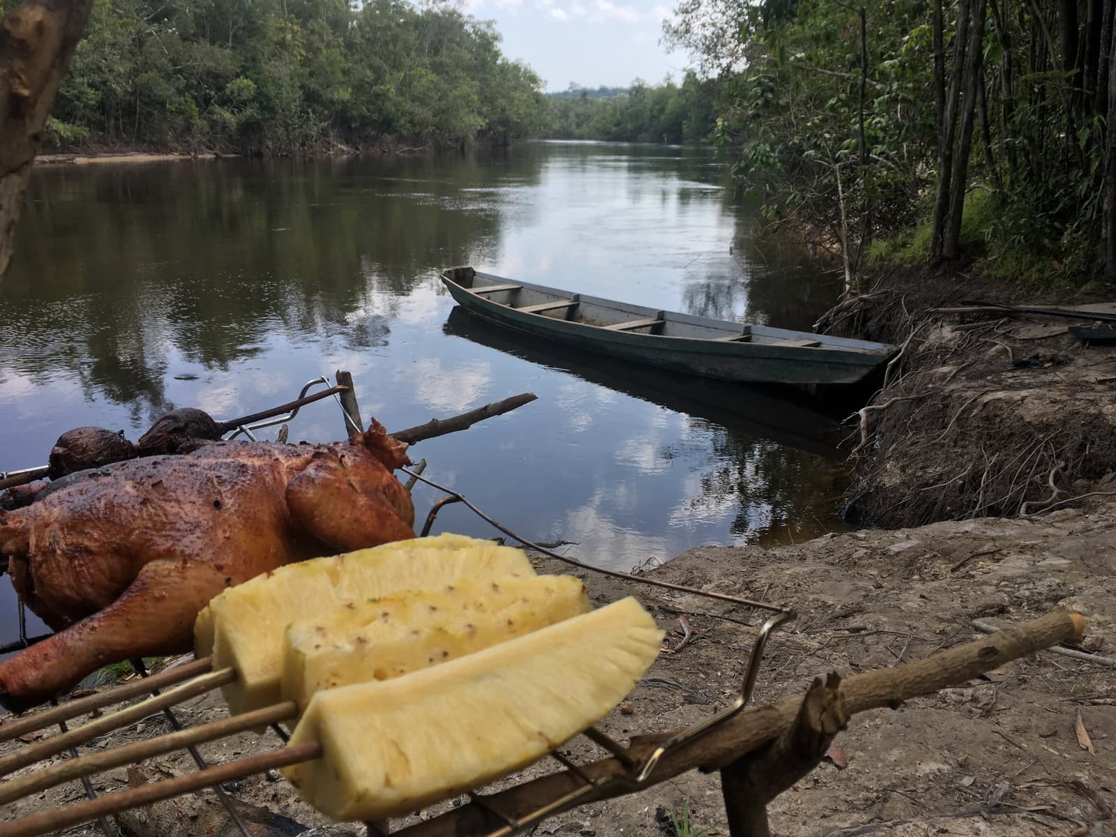 Lunch sorted, canoe ready, the Amazon sets its own pace. Grilled meat and fresh pineapple slices on bamboo skewers at the riverbank during an Amazon kayak tour in Brazil, with a wooden canoe moored on a calm jungle river behind