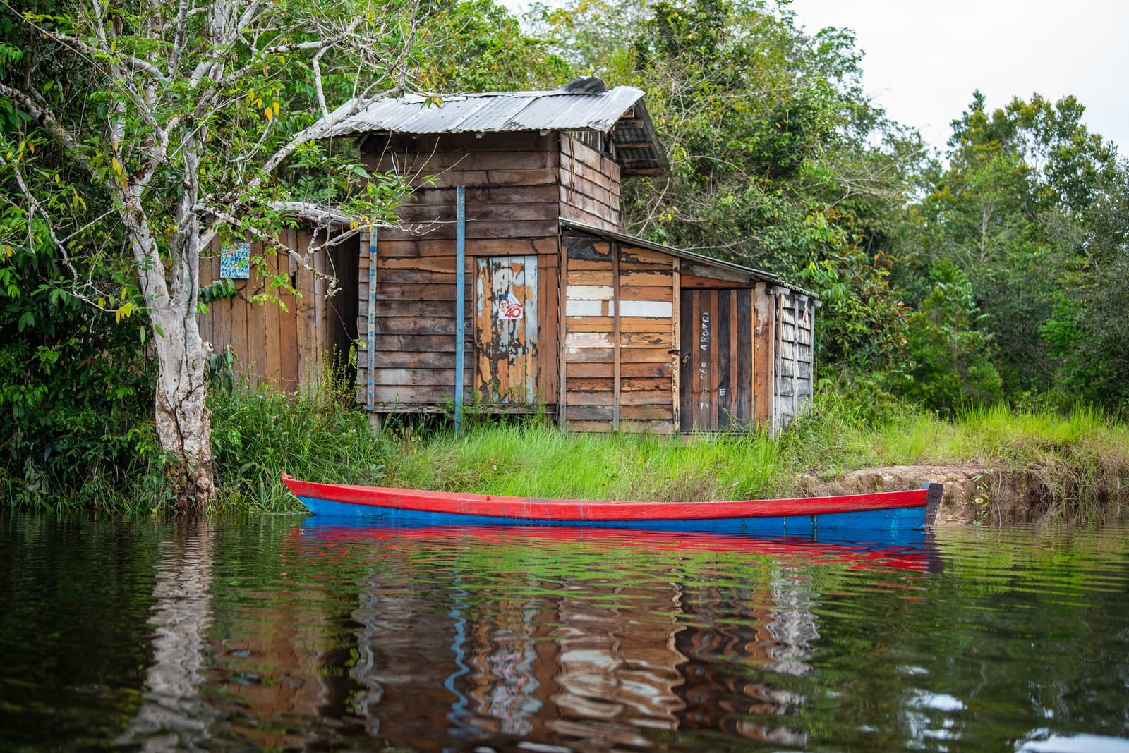 Life on the river runs at its own pace, and every plank tells a story. A red and blue wooden canoe moored on a dark river in front of a weathered timber hut surrounded by dense rainforest vegetation, seen on an Amazon kayak tour in Brazil