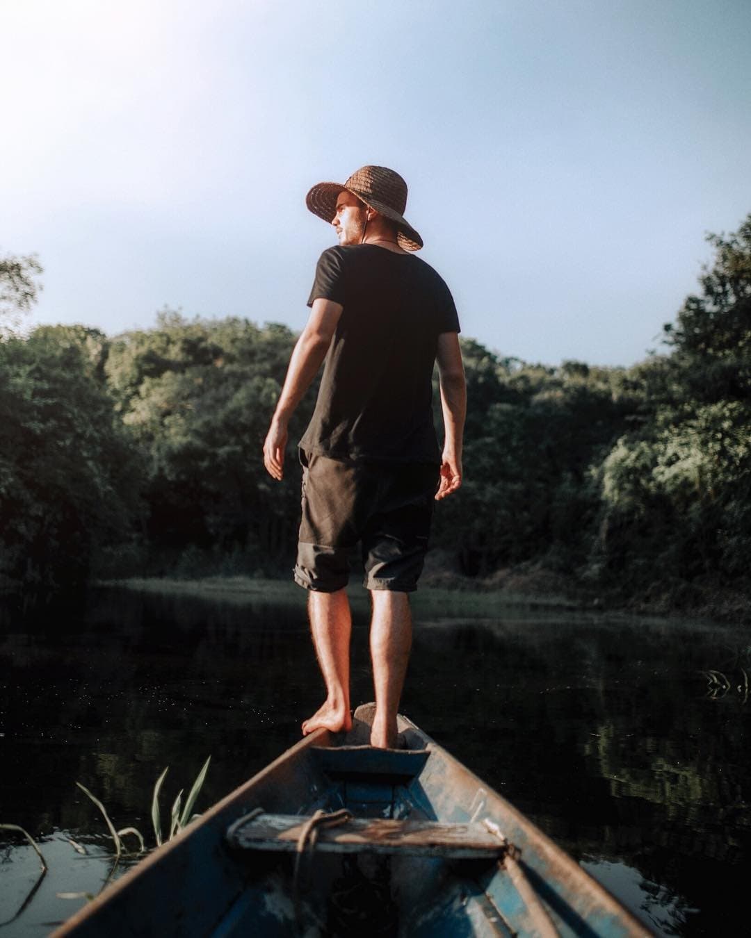 Bare feet, a borrowed hat, and a whole river ahead of you. A man in a straw hat stands barefoot on the bow of a weathered wooden canoe, gazing across a calm dark river flanked by dense green forest on an Amazon survival tour
