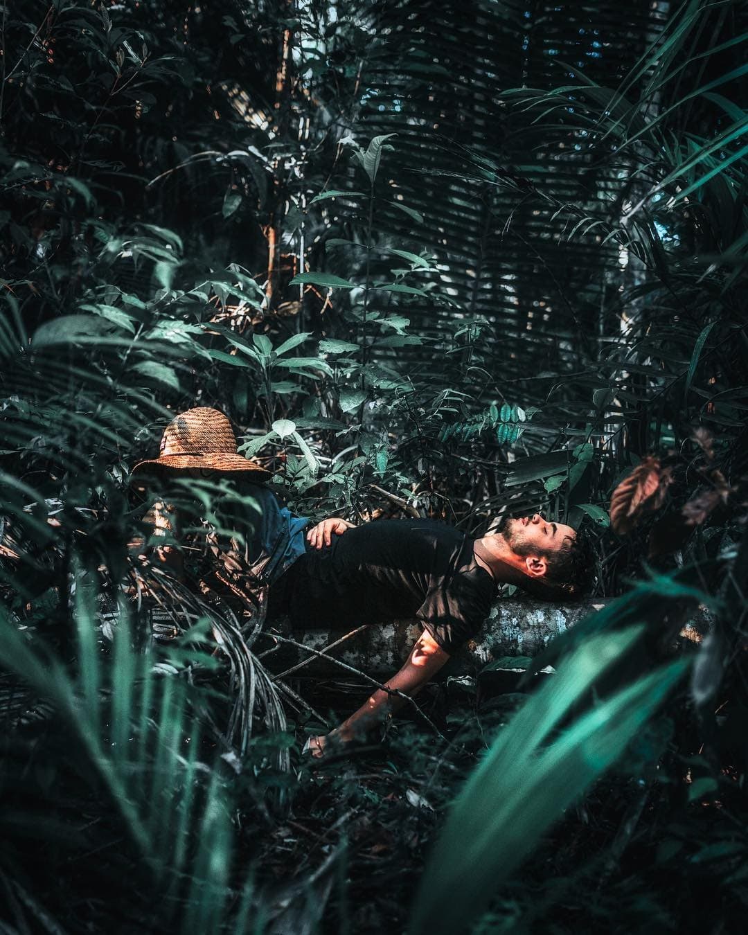 The forest has a way of making you stop. And then stay. A man reclining on a mossy log deep in dense rainforest undergrowth during an Amazon survival tour, his straw hat resting beside him amid layers of tropical leaves