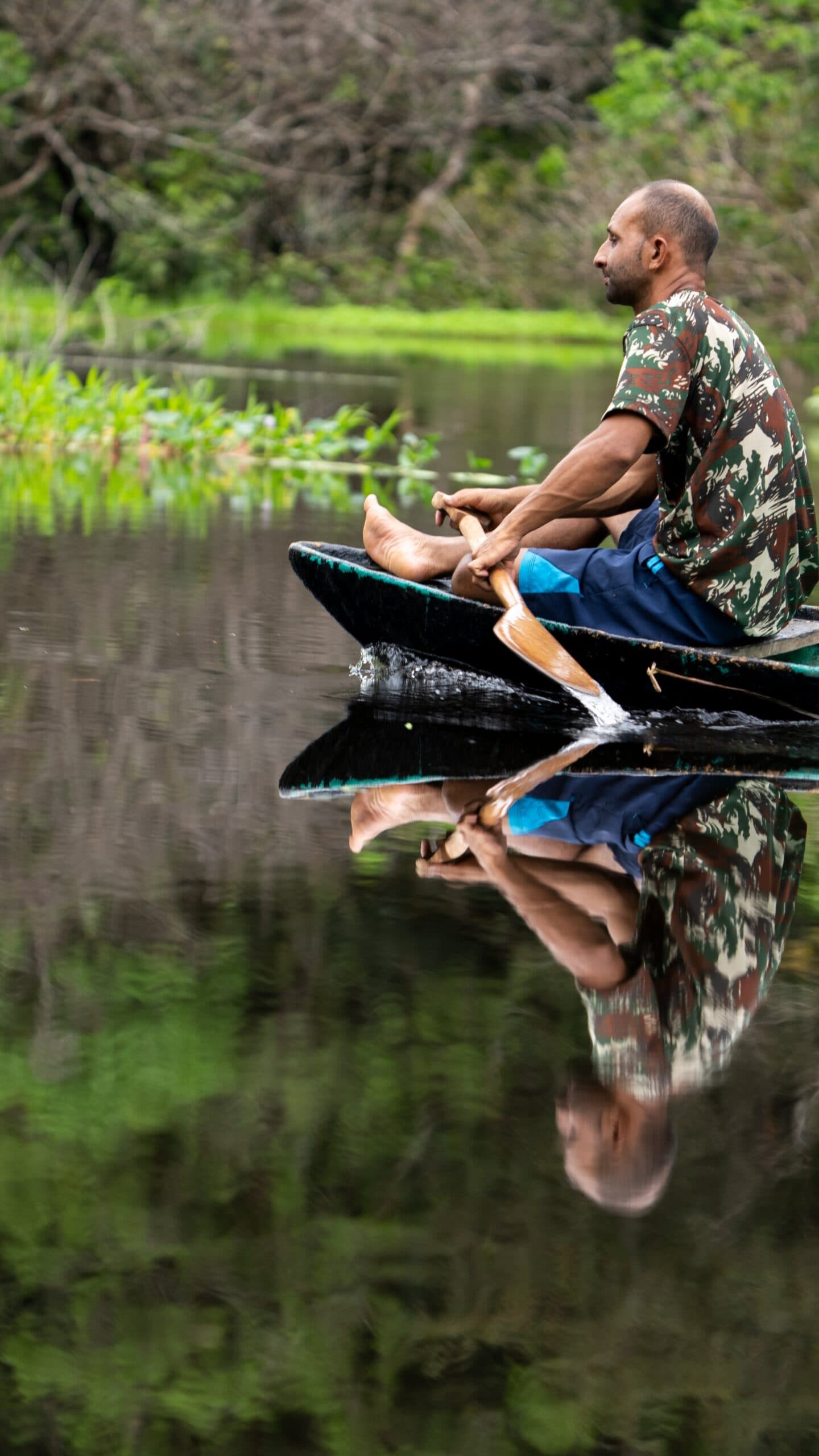He knows every bend of this river without a map. A man paddling a small wooden canoe on a glassy Amazon waterway, his perfect mirror reflection visible on the still dark water, with lush green vegetation along the bank — a quiet moment on an Amazon trek and survival tour