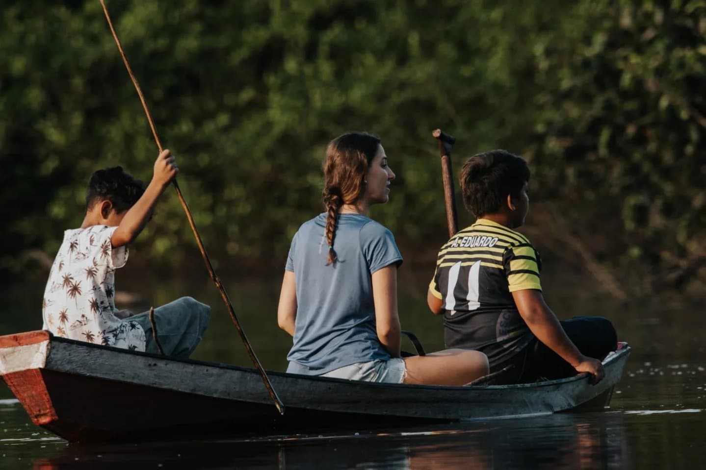 The boys know every bend in this river. She's catching up fast. A woman and two local boys paddle a wooden canoe along a calm jungle river during an Amazon trek and survival tour, dense green forest lining the banks