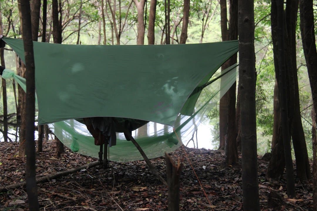 Your bed for the night, rigged and ready before the rain arrives. A green tarp and mosquito-net hammock rigged between trees in dense forest undergrowth, the jungle survival Brazil setup sheltering gear and bedding on a leaf-strewn forest floor